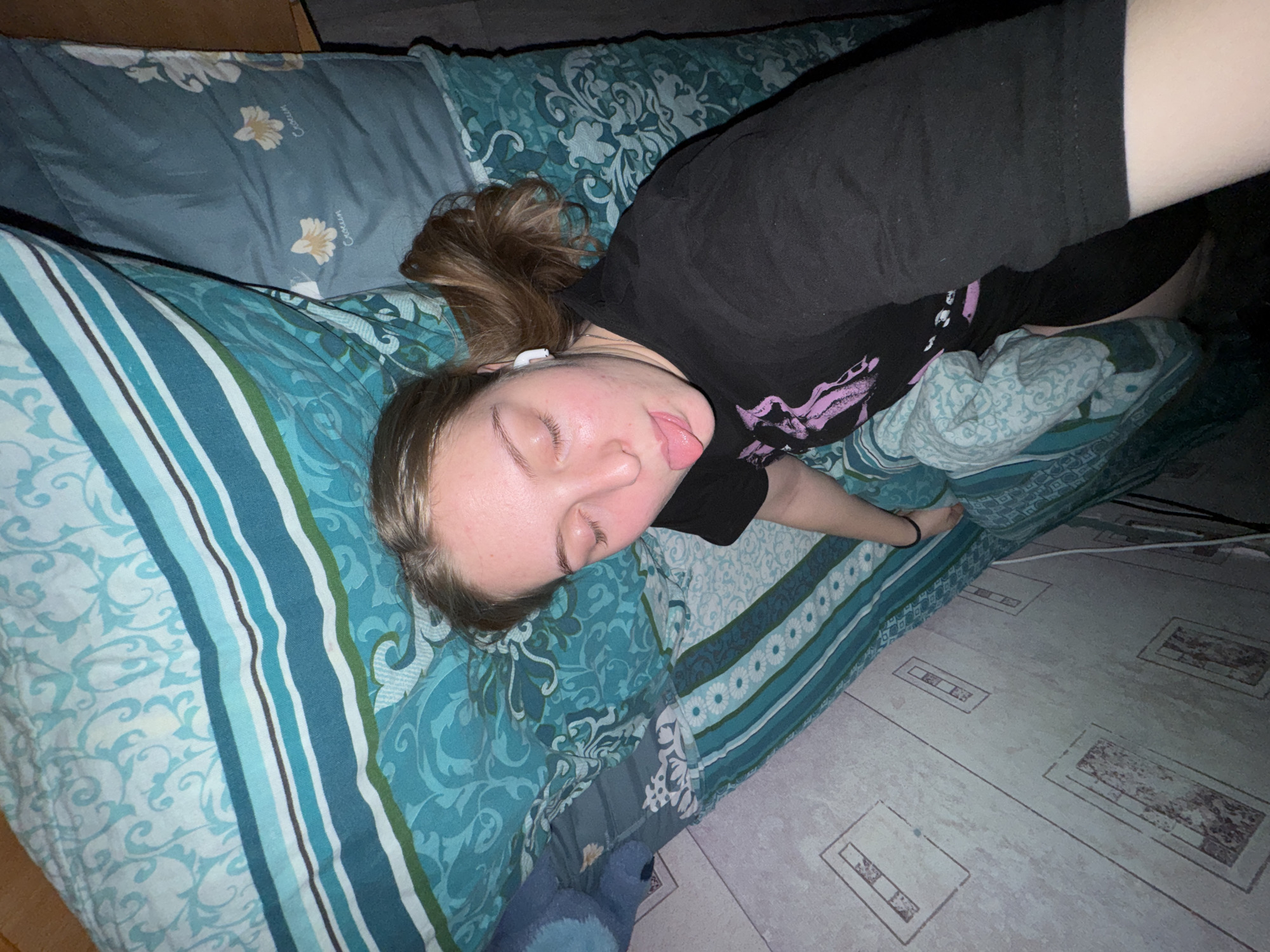 The image displays a young female lying on a bed in a bedroom in Perm, Russia. The female has light skin, brown hair, closed eyes, and her tongue is slightly extended, indicating a playful or relaxed pose, likely for a selfie. She is wearing a black short-sleeved top with a faint purple graphic design on the left shoulder and black shorts or leggings. A white wireless earbud is visible in her right ear.
The bed is covered with patterned bedding. The main duvet or comforter features horizontal stripes in shades of teal, blue, white, green, and brown, alongside a teal floral scroll pattern. A lighter teal sheet with a white floral design is visible beneath. Another section of the bedding, possibly a pillow or an additional blanket, is dark blue-grey with yellow and white floral motifs and the text "Crocus" partially visible. A blue plush object is situated on the bed in the upper right portion of the frame.
The ceiling above is light-colored and exhibits several rectangular panels, some with a subtle marbled or discolored appearance. A white cable runs along the wall area in the lower left section of the image. The perspective is from a low angle, consistent with a self-portrait taken while lying down.