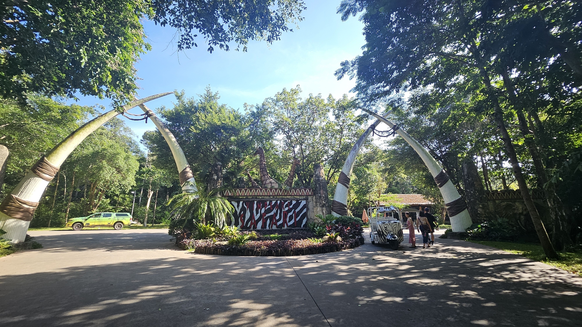 The image captures the entrance to a safari park, likely in Phú Quốc, Vietnam, given the presence of tropical foliage. The entrance is marked by two large, curved structures resembling tusks, wrapped with brown material. In the background, a decorative wall features a "SAFARI" sign with a zebra-stripe pattern and animal statues. To the left, a green safari vehicle is parked on a paved road. On the right, a smaller, zebra-striped vehicle is stopped, with two individuals standing near it. The scene is set amidst lush greenery, with tall trees and dense foliage forming a natural canopy, and the ground is paved with a dark asphalt material. The sky above is clear and blue.