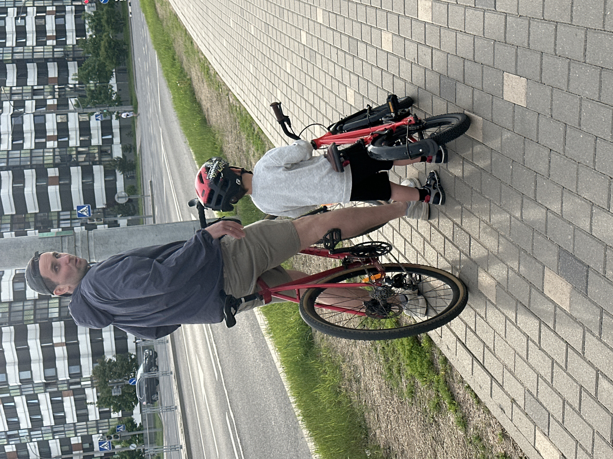 A man and a child are positioned next to a red bicycle on a paved sidewalk in Бараўлянскі сельскі Савет, Belarus. The man is mounting the bicycle, looking towards the camera. He is wearing a dark jacket over a light shirt, khaki shorts, and black shoes. A baseball cap is on his head. The child, wearing a gray sweatshirt, black shorts, and black sneakers, stands beside a smaller red bicycle lying on the ground. The child wears a helmet. The setting is outdoors with a road and a grassy area separating the sidewalk from the road. A large, modern building is in the background across the road.