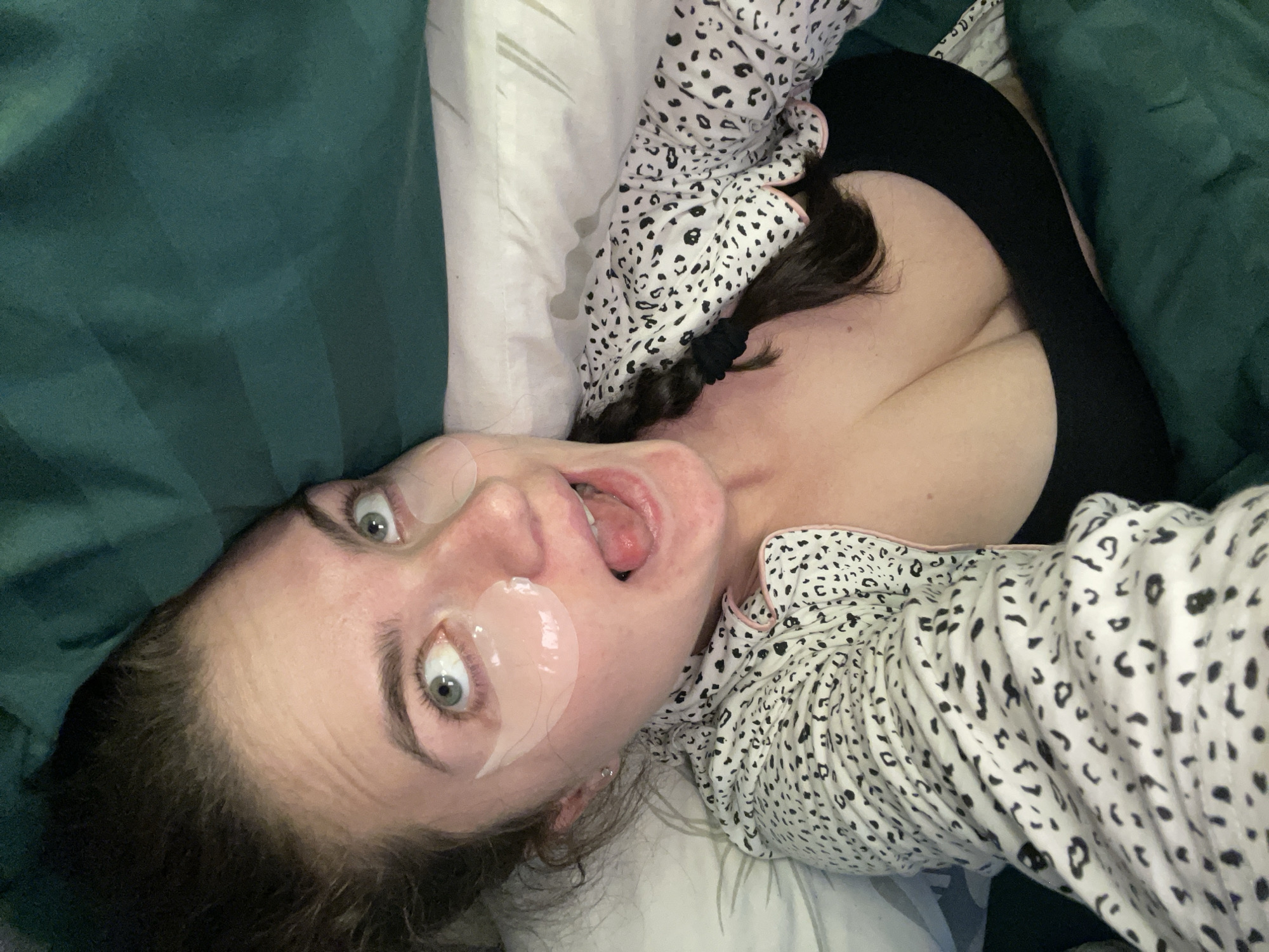 A woman is lying on a bed covered with green and white bedding, wearing a black tank top and a white pajama top with black spots. She has eye masks under her eyes and her mouth is open. The image does not provide any information to suggest that the location of the scene is in the city of Мосметаллоконструкция Онт, Russia.