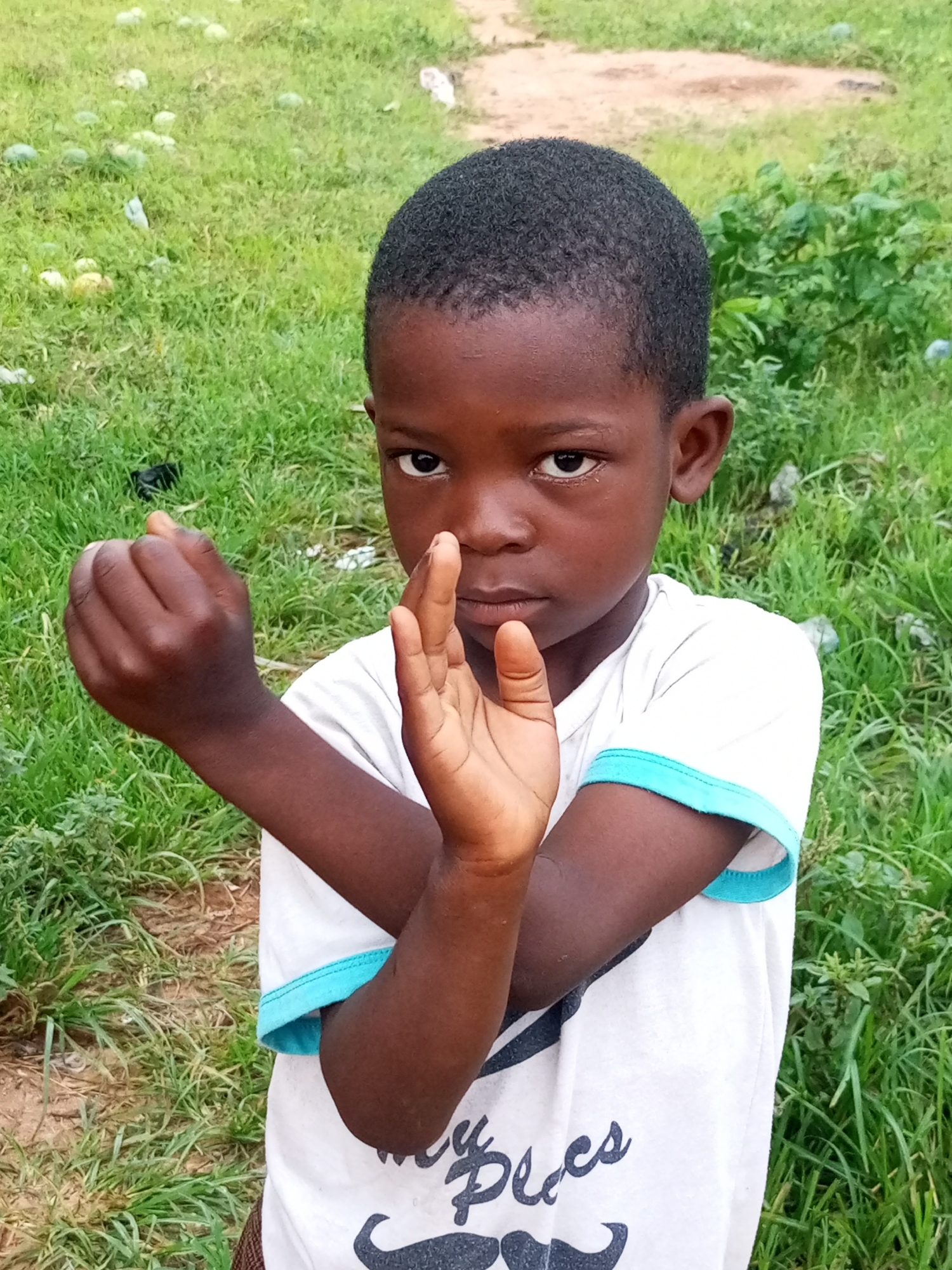 A young boy stands outside with a serious expression, his arms crossed in a defensive or martial arts pose. He is wearing a white t-shirt with blue trim around the collar and sleeves, and has short black hair. The background is a grassy area with some dirt patches and a few scattered objects, suggesting an outdoor, possibly rural or suburban, setting. There are no clear indicators of it being in Tempure, Nigeria.