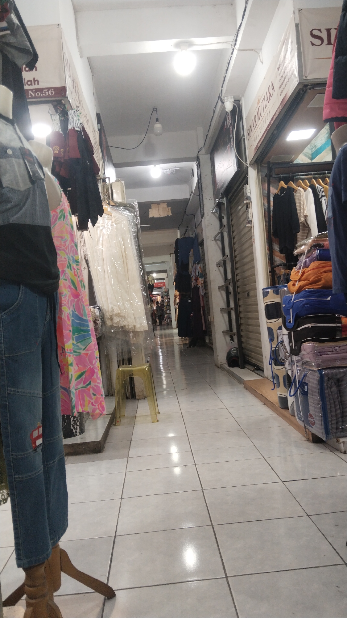 The image depicts a dimly lit indoor market aisle in Kamasan, Indonesia. The aisle is lined with shops on both sides, displaying clothing and other merchandise. The floor is tiled with large, light-colored squares, reflecting the overhead lighting. On the left side, a mannequin wearing blue jeans and a dark grey top is partially visible, along with a colorful, patterned dress. Further down the aisle, more clothing is displayed on racks and mannequins, some covered in plastic. On the right side of the aisle, shelves are stacked with folded textiles, possibly bedding or towels, in various colors. The shops themselves have metal roll-up doors, suggesting they are closed or preparing to close. In the distance, a few people are walking, indicating ongoing activity within the market. The overall impression is one of a bustling but somewhat cluttered retail environment.