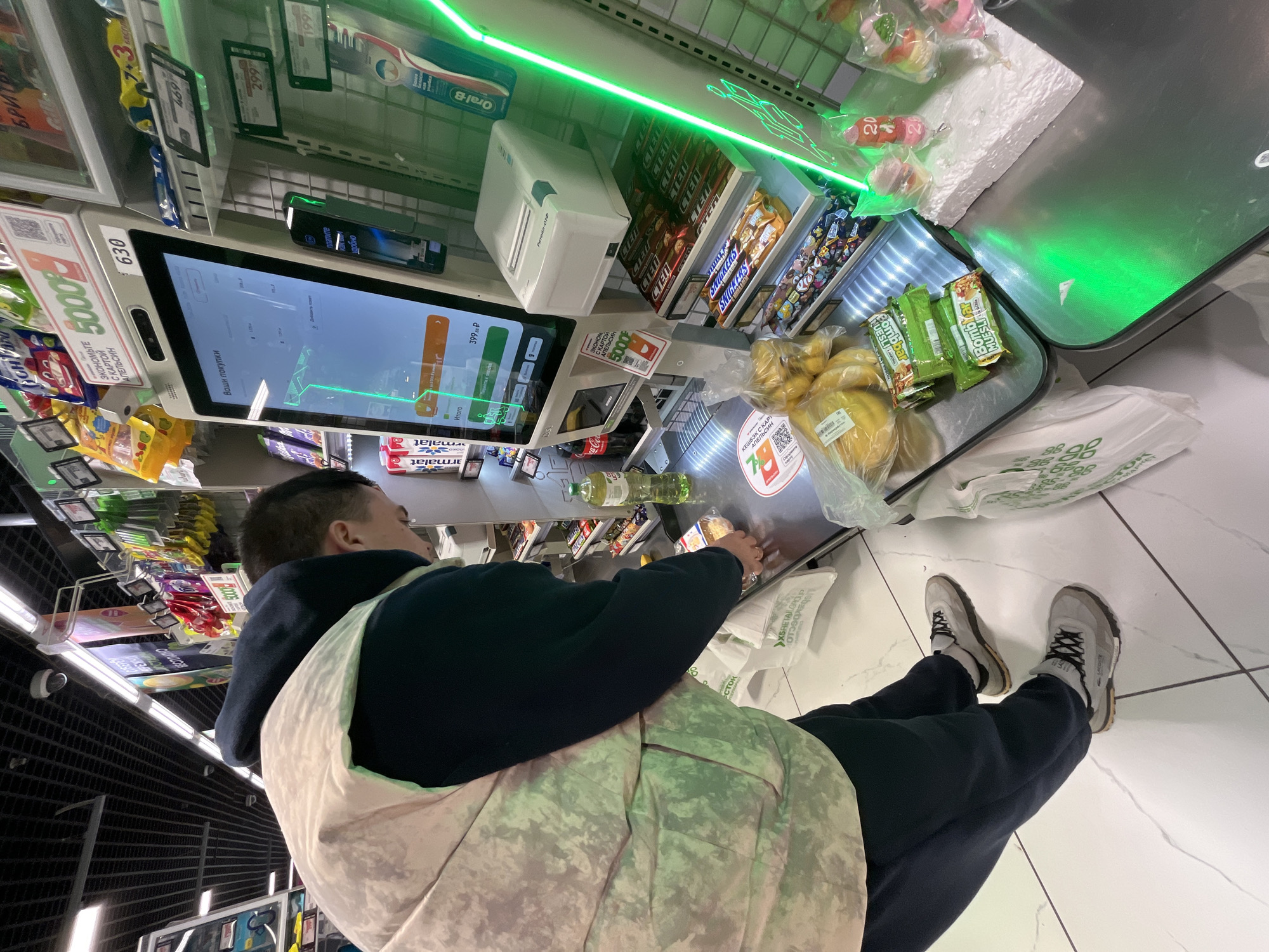 A high-angle shot shows a person using a self-checkout in what appears to be a supermarket. The person is wearing a dark hoodie, dark pants, and light-colored sneakers. A patterned bag hangs on their left shoulder. The self-checkout station displays a digital screen with purchase information in Russian, including the price "399 P" and the word "Итого" (Total). The screen is labeled "630" on its top left corner and "5000" and "экономьте с картой апельсин" (save with an orange card) on its bottom left corner.
A variety of items are in front of the person: bananas, granola bars, and a bottle of oil. Groceries are arranged in a small silver tray. The station has various items for sale, including candy, chocolate bars (Snickers, STEI), and Oral-B toothbrushes. Green neon lights are positioned above the area. Bags with Russian text are on the floor next to the checkout station. The background is a tiled floor. The OCR labels include "KESHBEK" (cashback), "MARKET", "ARMAT" (a brand of chocolate), and various price tags. From the text in Russian and the products on display, the location is likely a supermarket in Moscow, Russia.
