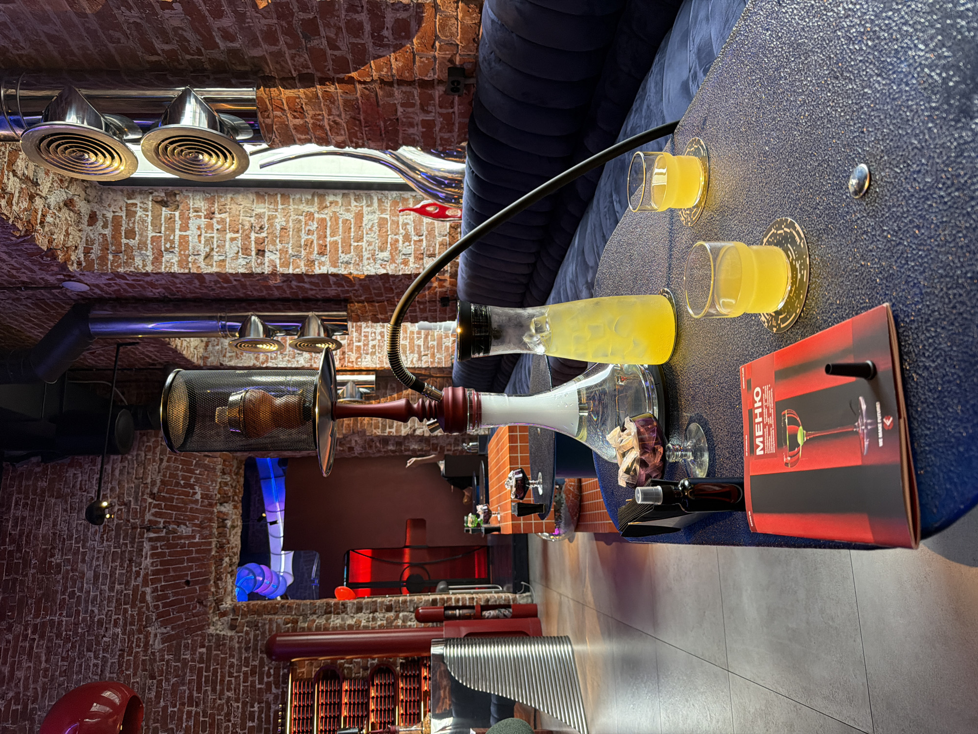 The image depicts the interior of a hookah lounge, likely in Moscow, Russia, characterized by its exposed brick walls, industrial-style ventilation systems, and modern, dimly lit ambiance. In the foreground, a blue, textured table displays a hookah with a white base and a red stem, a pitcher filled with a yellow beverage and ice, two small glasses of the same yellow beverage, a red menu booklet with the word "МЕНЮ" prominently displayed, and a small dark bottle. The background reveals a plush blue seating area along a brick wall, with further seating visible in the distance through an arched doorway. The lighting is atmospheric, with spotlights and colorful illumination from the background creating a moody atmosphere. There are no people clearly visible in the image, suggesting a quiet or early-hour scene.