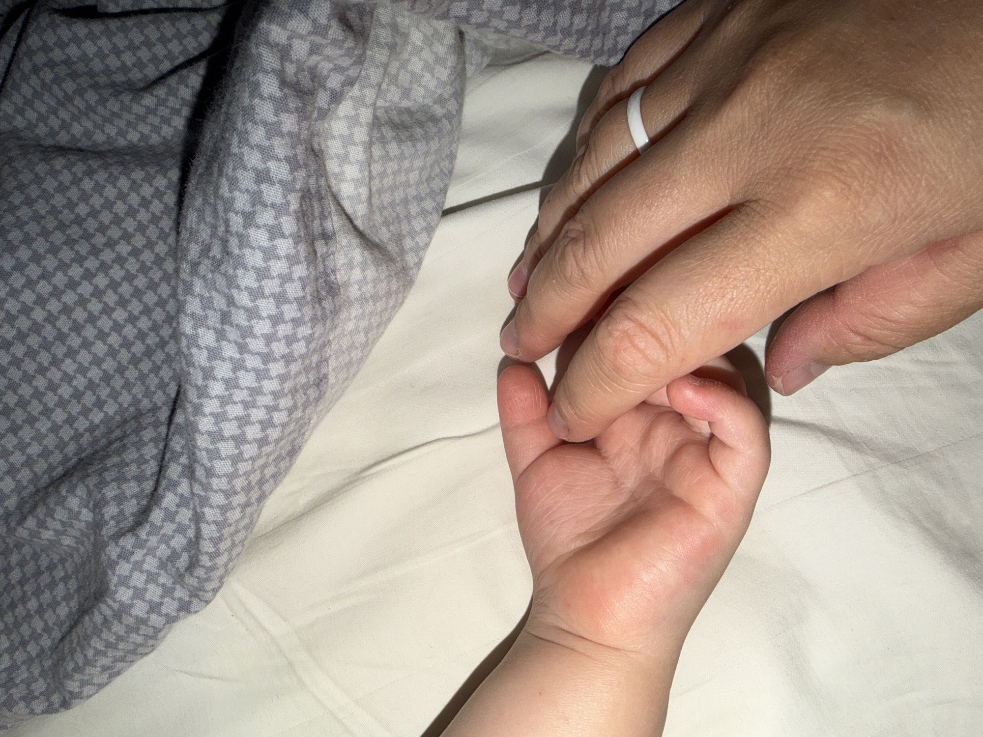 A close-up shot shows an adult's hand wearing a white ring, gently holding the hand of a baby. The hands rest on a white, unpatterned surface. A gray and white checkered fabric is visible on the left side of the image. The location of the image is not indicated.