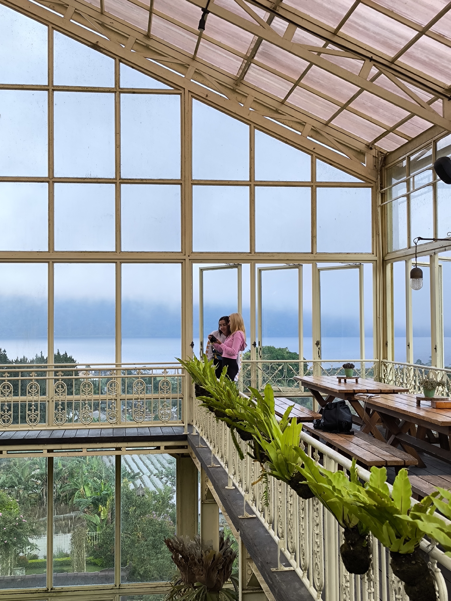 The image shows the interior of a greenhouse-like structure in Candikuning, Indonesia. The building has a metal frame with large glass panels. Two women stand on a mezzanine overlooking a lake or ocean obscured by fog. The woman on the right wears a pink sweater and holds a phone or camera. The woman beside her wears a dark top. A decorative metal railing with potted ferns runs along the edge of the mezzanine. Wooden picnic tables and benches are visible on the mezzanine. The lower level features a view of tropical plants through glass windows.