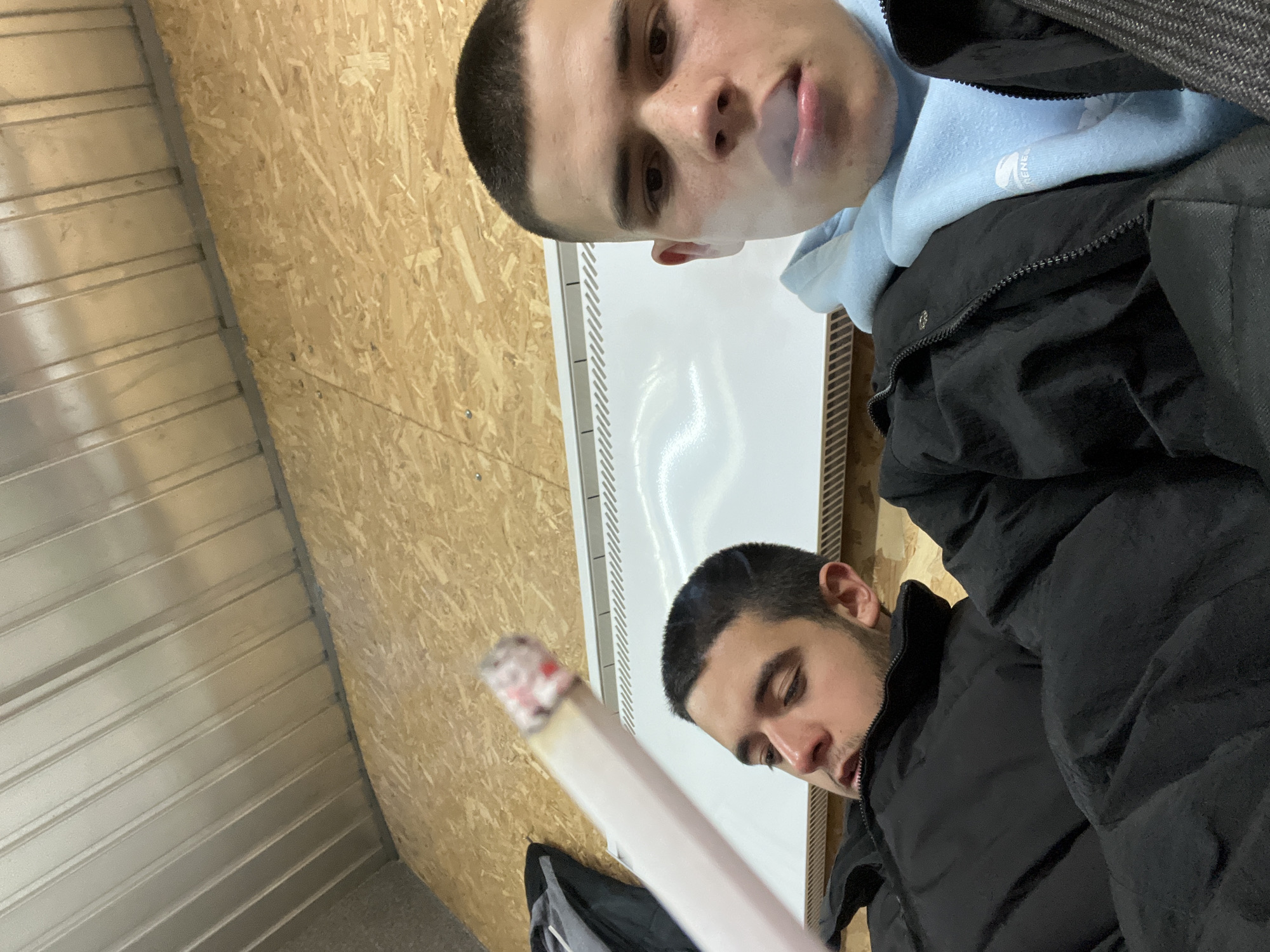 Two men are indoors. The man on top exhales smoke. He wears a light blue hooded sweatshirt under a black jacket. The other man holds a lit cigarette. He also wears a black jacket. A white radiator is behind them. The ceiling is corrugated metal and the walls are OSB. The location is unknown.