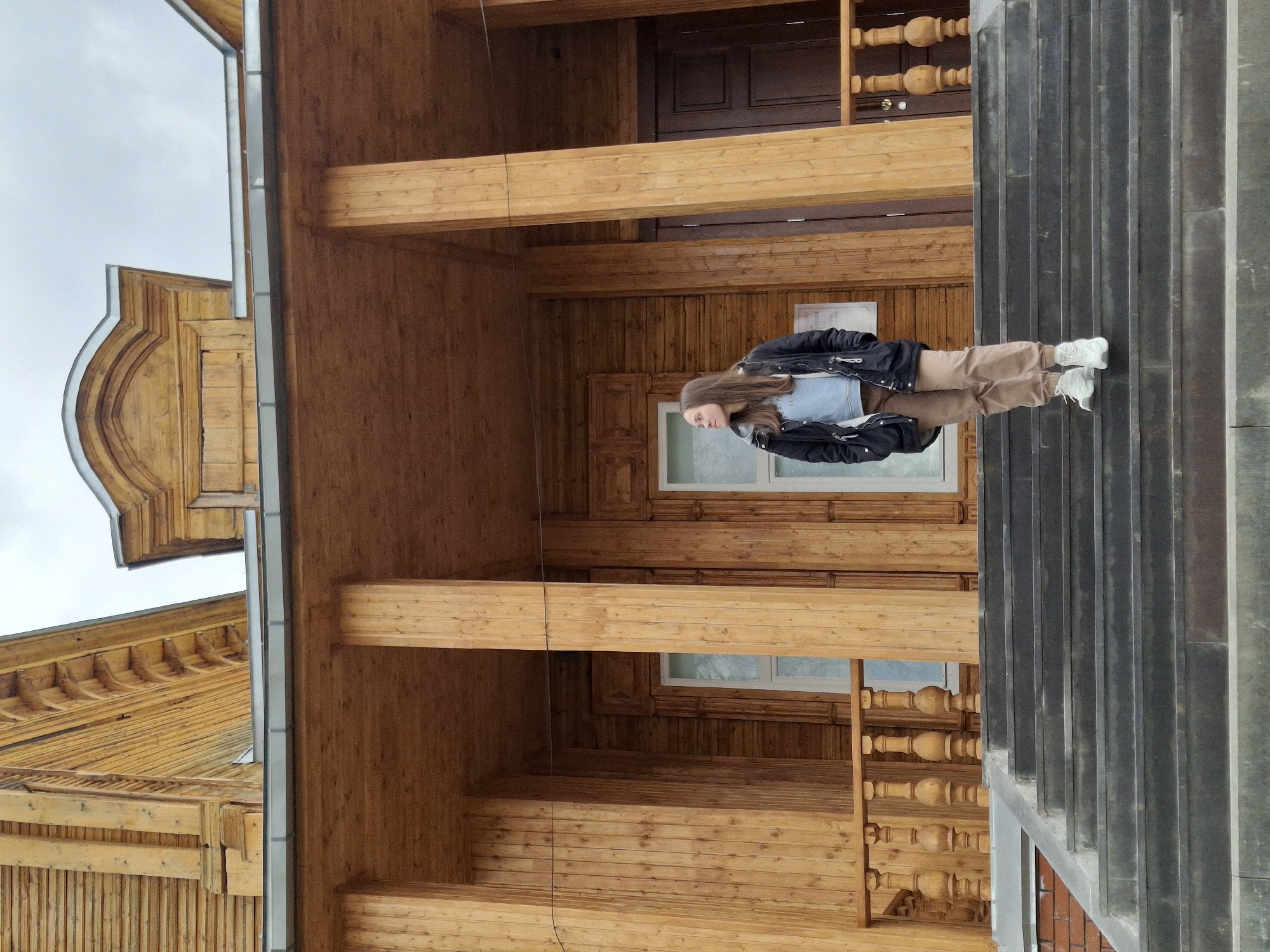 This media file depicts a young woman standing on a monumental staircase in front of a large, intricately designed wooden building, likely in Sarapul, Russia. The image was captured outdoors on an overcast day.
The central subject is a young woman, positioned slightly to the right of the frame, facing slightly left. She has long, dark brown hair and is dressed in a black puffer jacket over a light blue top, paired with light brown cargo pants and white sneakers. Her expression is calm and neutral.
The dominant background feature is a multi-story wooden building constructed with horizontal planks and large timber beams, showcasing a warm, rich wooden tone. The architecture suggests a traditional style, possibly a historical Russian log house or a grand timber structure. Notable details include decorative elements around a visible window and door, substantial wooden pillars supporting an upper level, and an upper balcony or porch with turned wooden balusters or spindles. A distinctive, elaborately carved wooden pediment or gable is visible on the roofline to the left.
In the immediate foreground, to the right, is a wide staircase made of dark, possibly stone or concrete, material. The steps have a textured, vertically ribbed or fluted pattern. The woman is standing on this staircase, which provides a stark material and color contrast to the wooden building behind her.
The sky is uniformly overcast, indicating a cloudy daytime. A small, light-colored rectangular sign with unreadable dark text is visible on the wooden wall behind the woman, next to a window. The overall scene captures a posed portrait against a backdrop of impressive traditional architecture.
