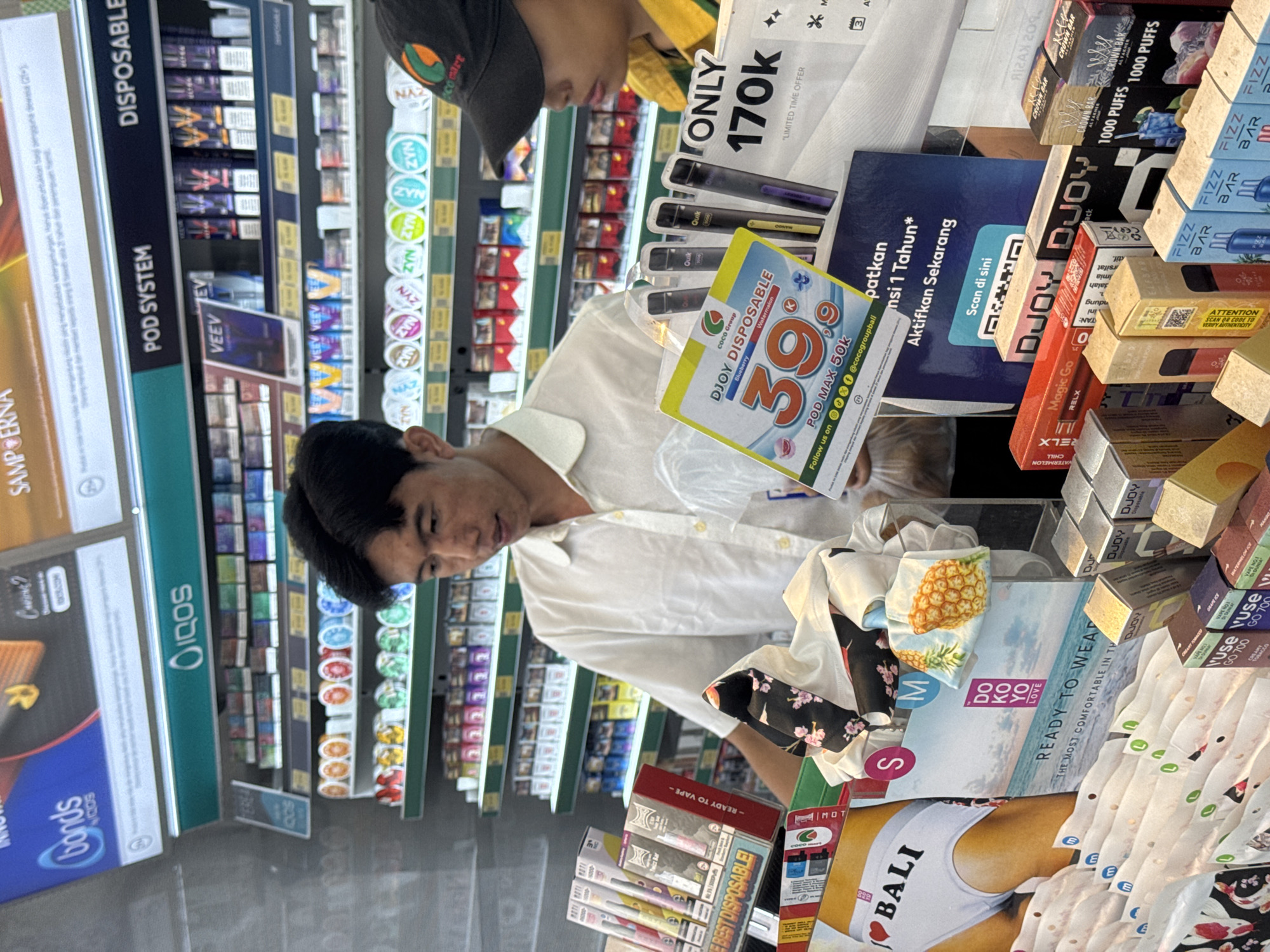 In a retail store in Kuta Selatan, Indonesia, a young man wearing a white short-sleeved shirt is standing behind a display of vape products. He is holding a plastic bag and a promotional flyer for "DJOY DISPOSABLE" vapes, which features a large "39" and mentions "POD MAX 50K". To his right, another person wearing a black baseball cap and a yellow and green shirt is visible. The shelves behind them are stocked with various vape brands and smoking accessories, including IQOS products, VEEV devices, and ZYN nicotine pouches. In the foreground, a display features underwear with "I ❤️ BALI" printed on it, as well as other vape products in their packaging. The store appears to be well-lit, with bright overhead lighting.