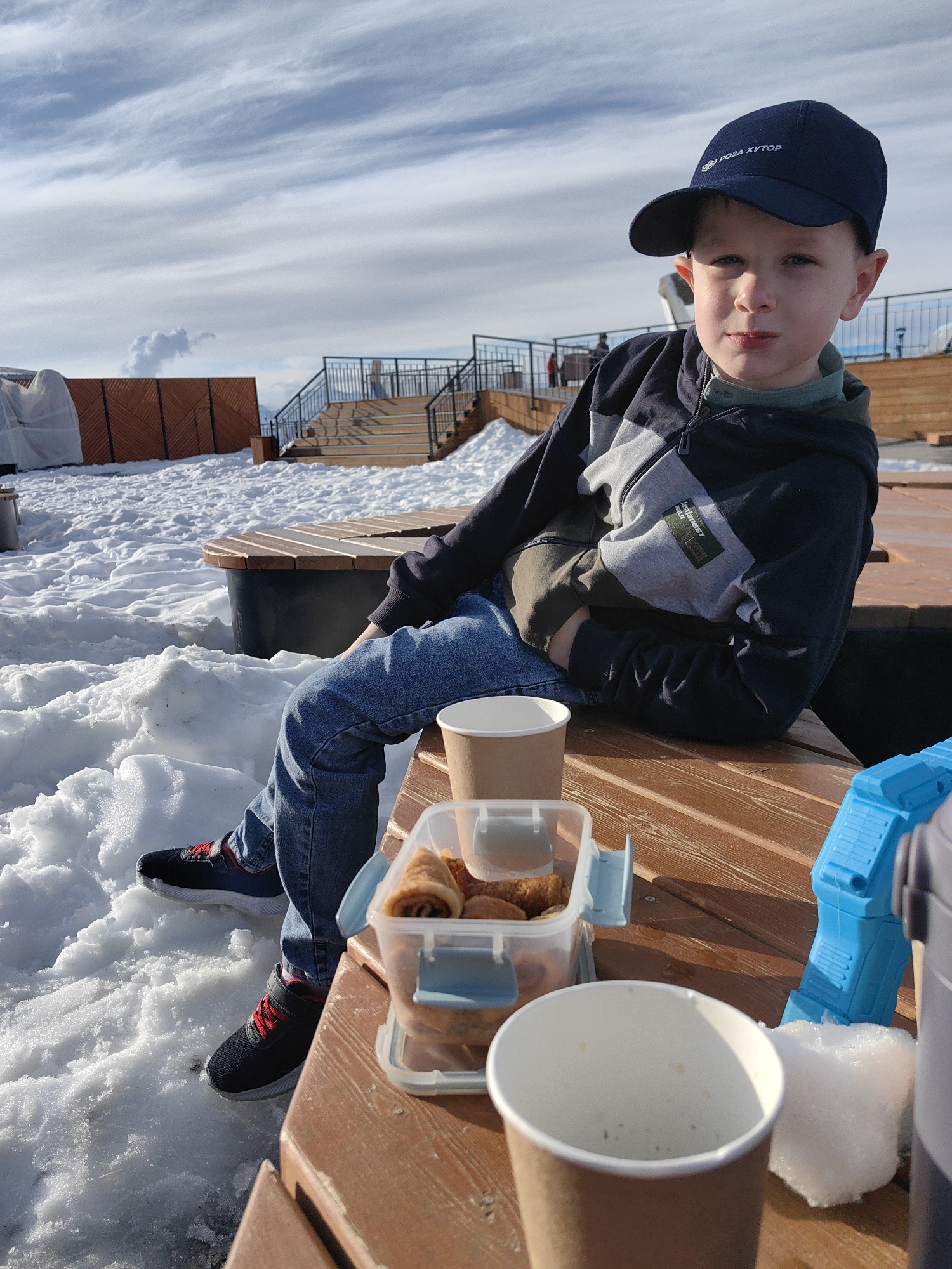 A young boy, approximately 6-8 years old, is seated on a wooden bench or platform in an outdoor, snowy environment, likely a winter resort in Estosadok, Russia, such as Rosa Khutor. He looks directly at the camera with a neutral expression.
The boy is wearing a dark blue baseball cap with the white Russian text "РОЗА ХУТОР" (ROZA KHUTOR) visible on the front. He has on a dark blue and grey zippered hoodie, with a small green patch on the sleeve that includes "bestfriend!" and "daily". Underneath, a green long-sleeve shirt is visible. He is dressed in blue jeans and black sneakers with red laces.
On the wooden surface in front of him are several items: two paper cups (one brown, one white and seemingly empty), and a clear plastic container holding food that appears to be rolled pancakes or crepes. Part of a blue plastic toy, possibly a snowball maker, is also visible to the right.
The setting is bright daylight, indicated by the clear lighting and visible shadows. The ground is covered in white, undisturbed snow, suggesting cold, winter weather. In the background, there are more wooden structures, including benches, a fence, and a staircase with black railings, typical of a ski resort. The sky above is light blue with sparse, wispy white clouds. The overall scene captures a moment of rest or a snack break during a day out in the snow.
