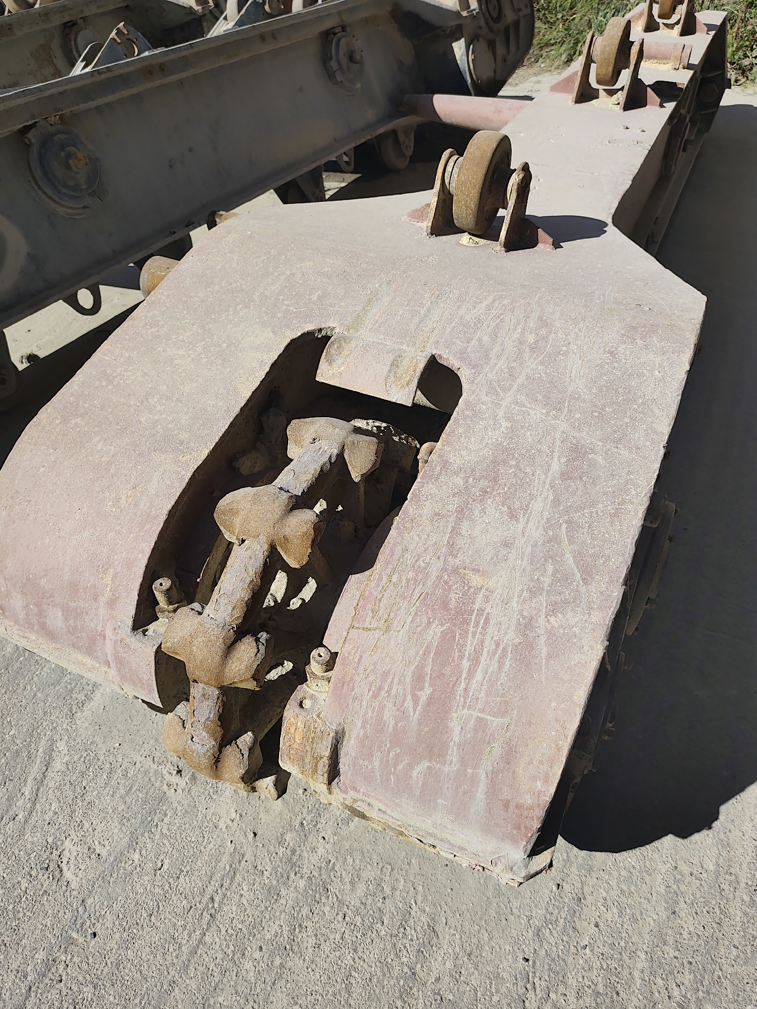 The image displays a close-up, high-angle view of heavy machinery, specifically the tracks and connecting components of what appears to be a large vehicle or construction equipment. The machinery is predominantly colored in a faded red and rusty brown, showing signs of age and wear. One prominent section is a wide, flat metal plate with a large, curved cutout revealing a complex assembly of interlocking metal parts that resemble a track or chain mechanism. This mechanism is also rusted. Mounted on top of the red plate are two U-shaped metal brackets, each holding a small, rusty wheel or roller. To the left, another section of heavy machinery, darker in color (likely gray or black), is visible, with visible bolts and circular fasteners. The machinery rests on a concrete or asphalt surface, and a shadow of one of the components falls across the ground. In the background, blurred green foliage suggests an outdoor, possibly industrial or construction site setting. The overall impression is one of disuse and weathering. There are no people or discernible city landmarks present in the image.