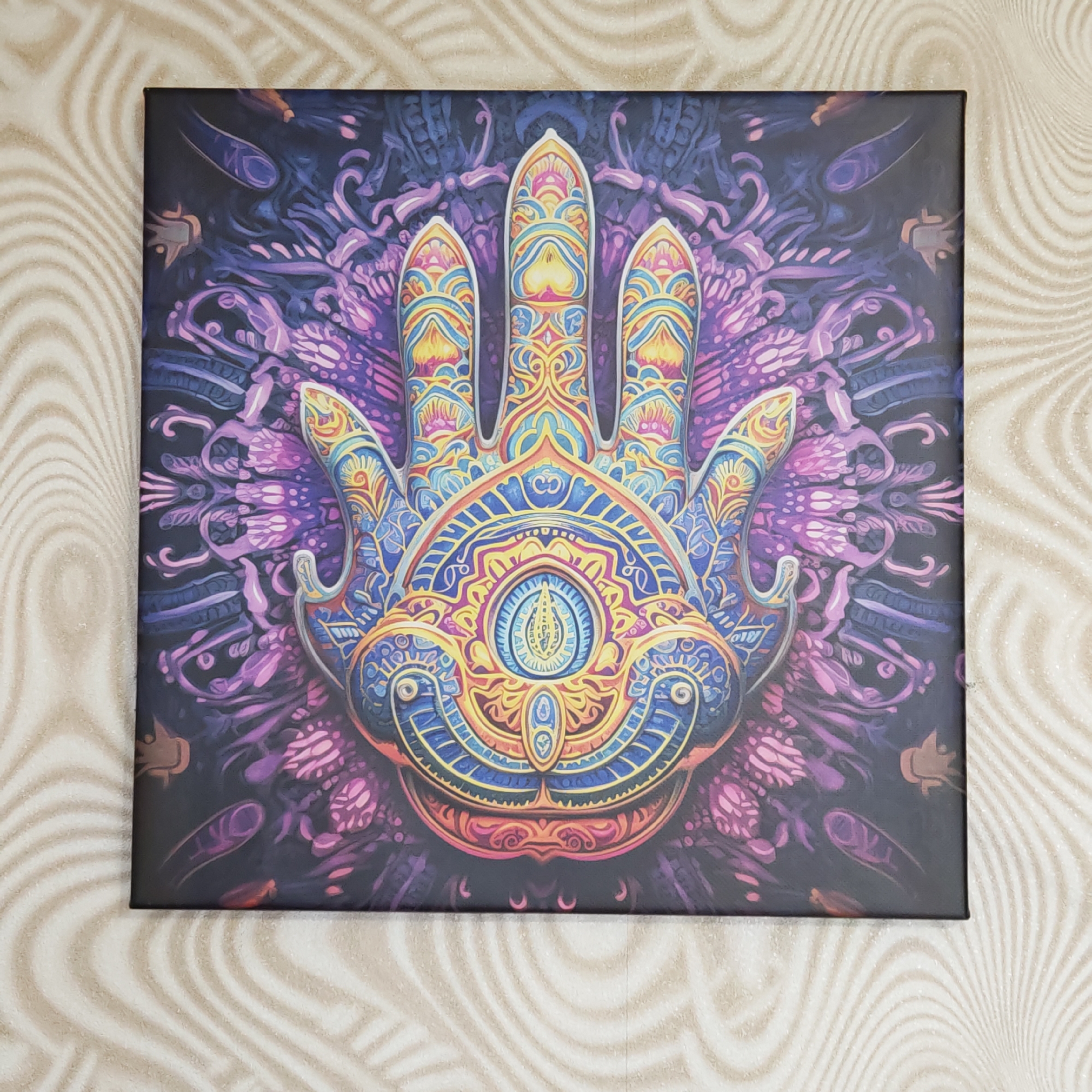 A square canvas print depicting a stylized, colorful hand with an eye in the palm is mounted on a wall covered in a swirling, beige and white patterned wallpaper. The artwork features a central hand motif with three elongated fingers, each elaborately decorated with yellow, blue, and orange patterns. The palm contains a detailed eye design surrounded by concentric circles of blue, yellow, and orange. The background of the artwork is a dark purple and blue, filled with stylized floral and geometric designs. The canvas has a black frame and is positioned against the wallpaper, which exhibits a subtle, wave-like pattern.
This image was NOT taken in the city of Ketoshi, Russia.