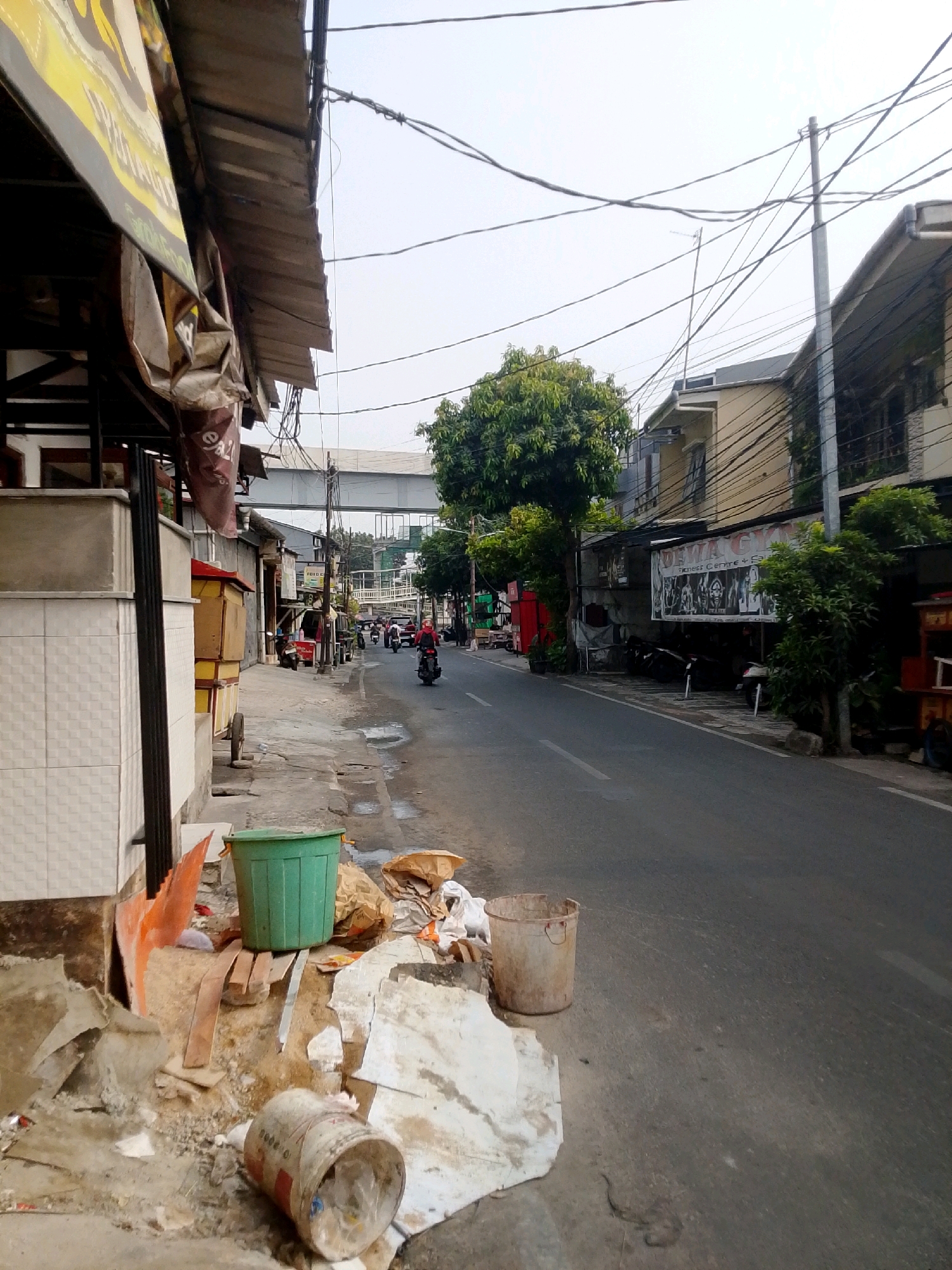 The image depicts a street scene in East Jakarta, Indonesia, characterized by a moderately busy road with some traffic, primarily motorcycles. On the left side of the street, a sidewalk is lined with various structures, including a building with a tiled facade, awnings, and what appears to be small businesses or stalls. Debris is scattered along the sidewalk, including construction materials, trash bags, and buckets, indicating some ongoing work or accumulation of refuse. Further down the street, trees line the sidewalk and the road, with more buildings visible in the background. A pedestrian bridge spans the road in the distance. On the right side of the road, a row of buildings, some with advertisements, are visible. A few motorcycles are parked along the sidewalk. The sky is overcast.