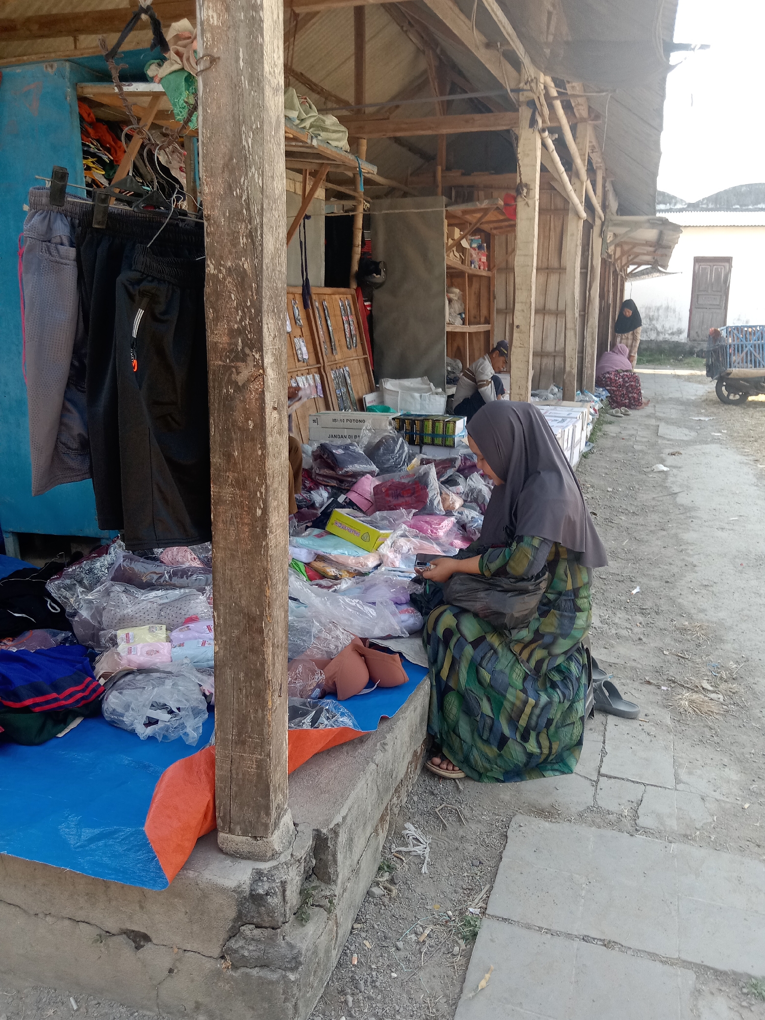 This image depicts a street market scene in Klepek, Indonesia. The foreground is dominated by a concrete stall with a rough wooden pillar. Several people are present: a woman wearing a dark grey hijab and a green, black, and blue patterned dress is seated on a dark object, looking down at what appears to be a mobile phone, with her feet in sandals. To the left, a blue tarp is spread out, laden with packaged clothing, including items in clear plastic. Hanging to the left of the pillar are several pairs of trousers. In the mid-ground, other individuals are visible: a person in a grey shirt and blue cap is seated, and another person in a light-colored hijab and patterned skirt is standing. Behind them, stalls made of wood and corrugated metal house various goods, including more packaged clothing and what appears to be canned beverages on shelves. The ground is a mix of dirt and concrete tiles. In the background, a white building with a wooden door is visible, along with a blue wheeled cart. The lighting suggests it is daytime, with bright sunlight casting shadows.