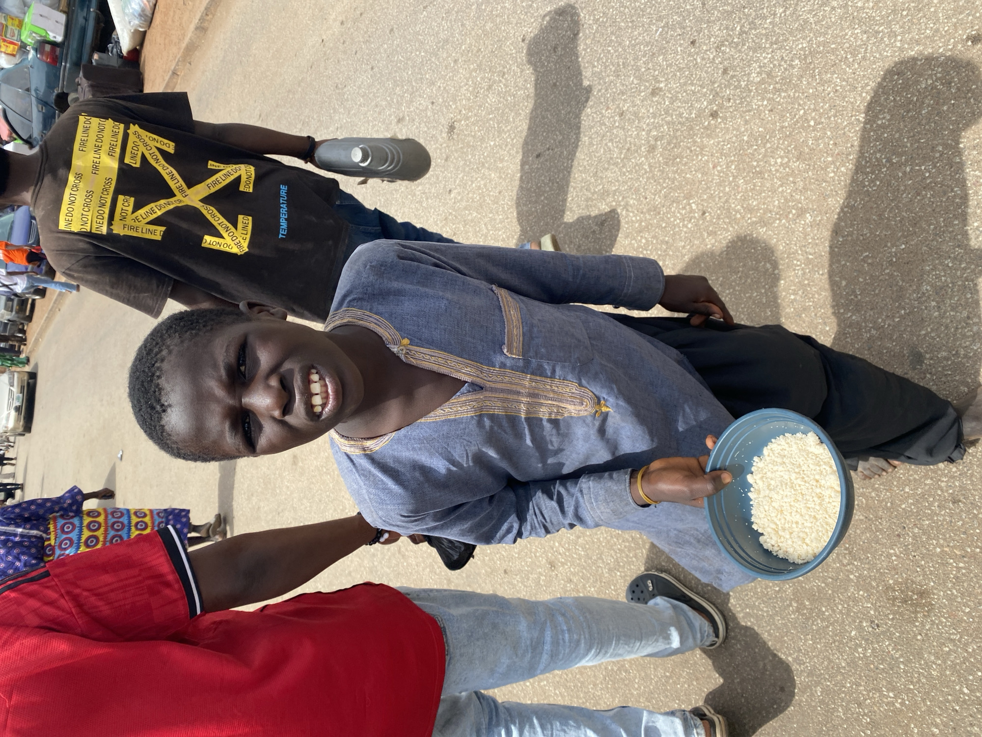 The image shows a young person holding a blue bowl of rice, possibly for sale or alms. The person is wearing a blue, long-sleeved shirt with gold trim. There are two other people in the frame, one wearing a red shirt and light-colored jeans, and another wearing a brown t-shirt with "FIRE LINE DO NOT CROSS" printed on it. The location appears to be a street in Jos, Nigeria, based on the appearance of the people and surrounding environment. The background shows parked cars and buildings. The ground is paved. The weather seems bright and sunny, judging by the shadows.