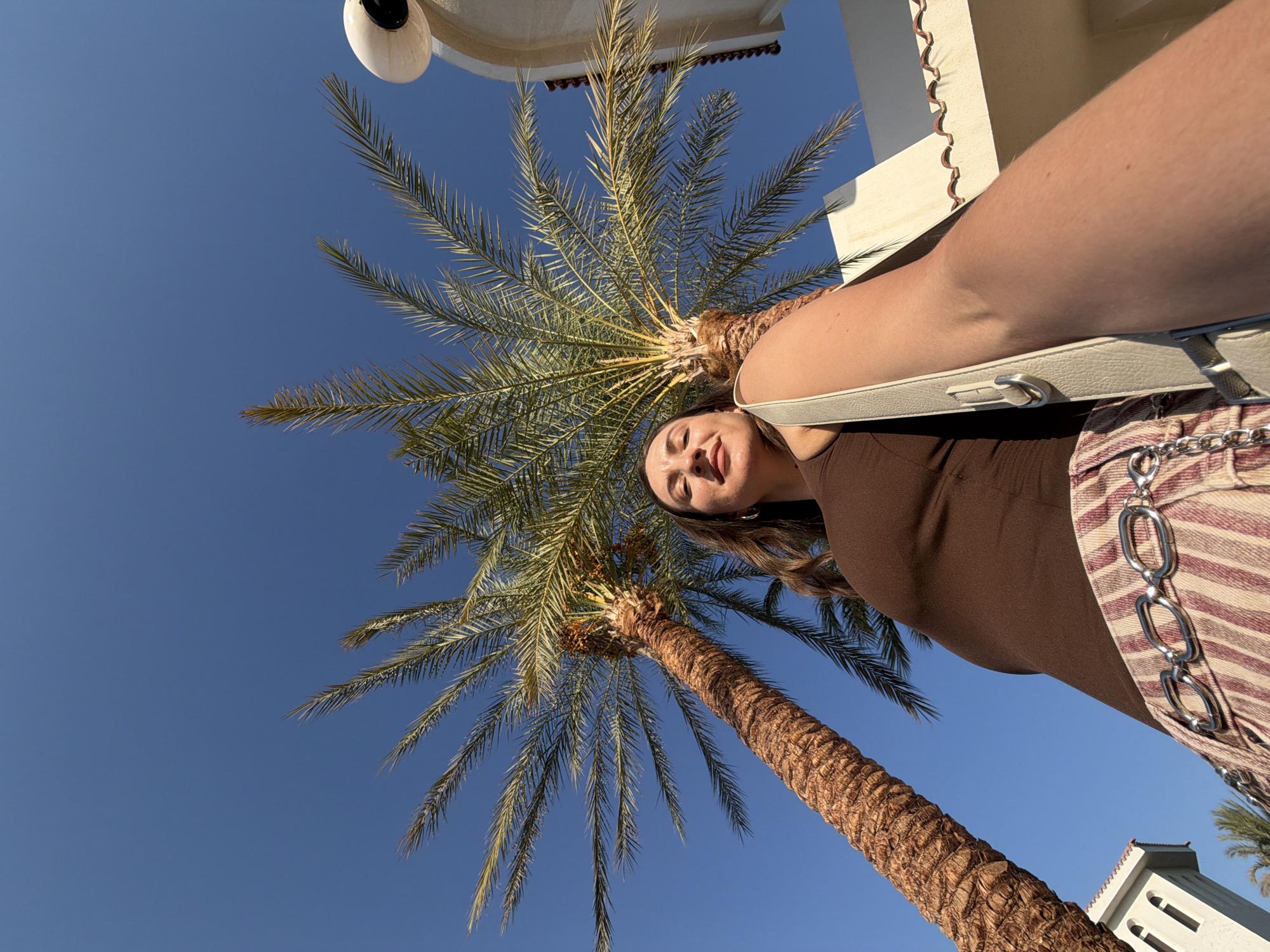 A low-angle, outdoor, full-body shot features a young Caucasian woman standing beneath two palm trees against a clear blue sky. She is looking up and slightly to her left, with her eyes closed and a gentle smile on her face. Her brown hair is down and falls over her shoulders. She is wearing a brown tank top and striped pants with a silver chain belt. A light-colored purse is slung over her right shoulder, with the strap extending across the front of her body. In the background, a white building with arched windows and a tiled roof is visible to the right. The scene is likely located in Sharm El Sheikh, Egypt, given the presence of palm trees and the architectural style. The sunlight is bright, casting shadows and highlighting the textures of the palm tree trunks and the woman's skin.