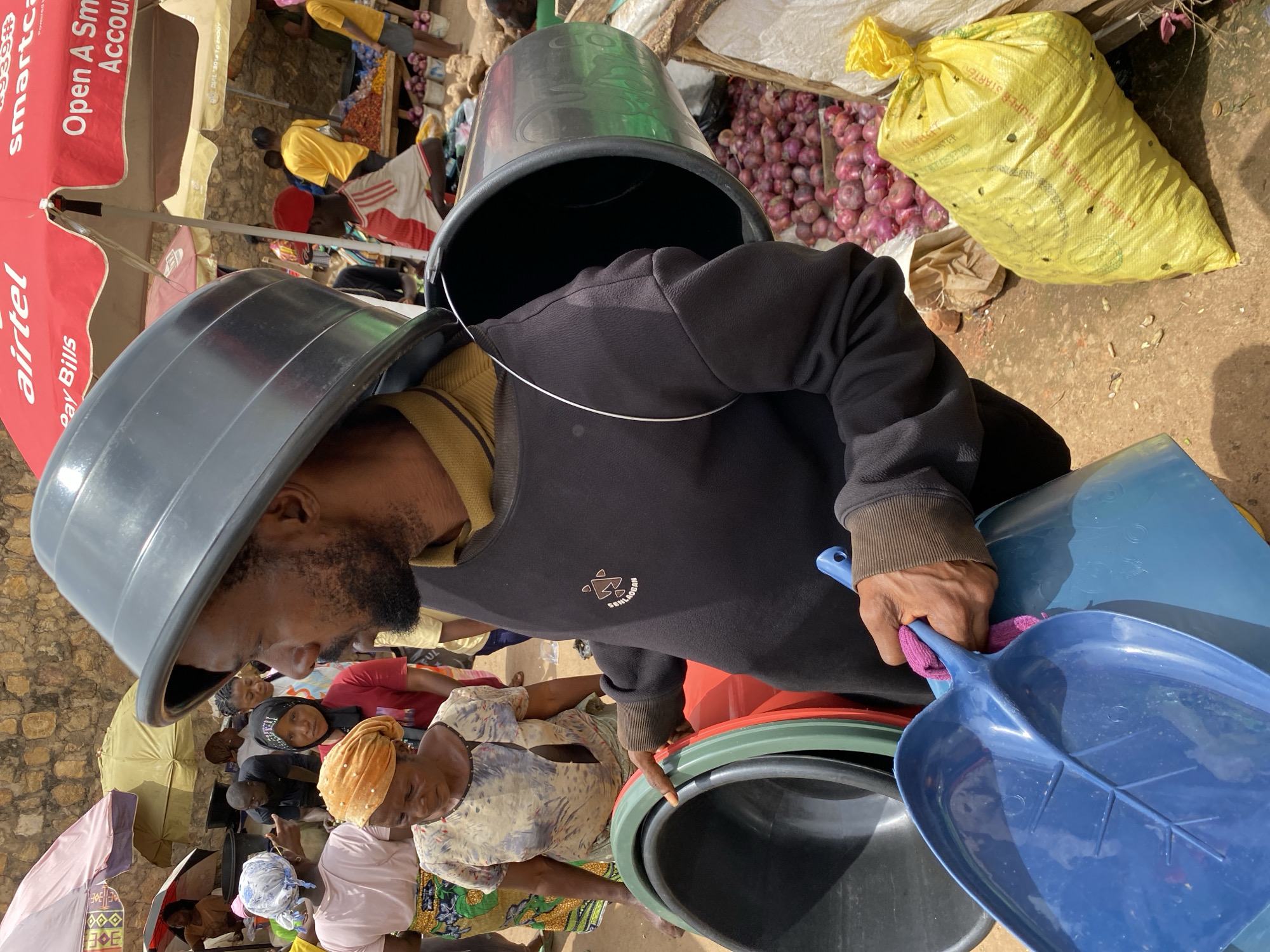 A man with a beard and dark skin is carrying large plastic basins on his head and in his arms. He is wearing a black sweatshirt with a yellow collar and dark pants. He is holding a blue plastic shovel in his right hand. In the background, there are people, market stalls, and umbrellas. One umbrella is red and has "airtel" written on it. There is a pile of red onions next to a yellow plastic bag. The scene appears to be a busy market in Bukuru, Nigeria.