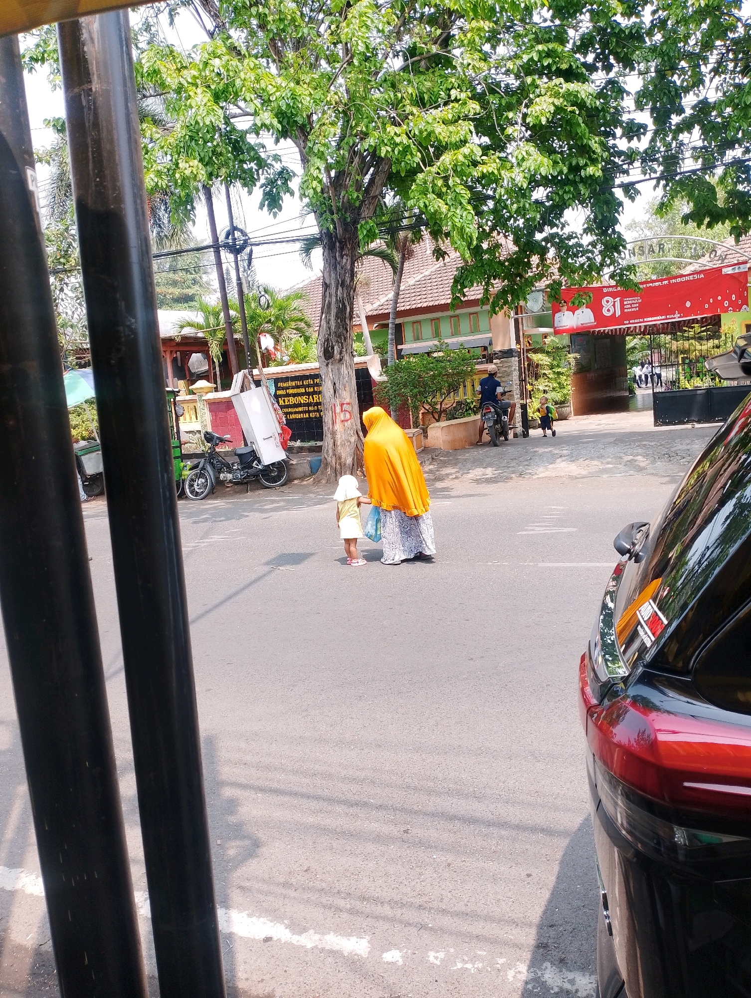 In Probolinggo, Indonesia, a street scene unfolds under a bright sky. Two people, a woman and a child, stand on the paved road, partially obscured by dark vertical poles in the foreground. The woman, wearing a vibrant orange headscarf and a patterned skirt, holds a plastic bag. The child, in a light-colored dress and head covering, faces away from the viewer.
Behind them, a black motorcycle is parked on the side of the road, carrying a large white box on its rear. A sign is visible on a building in the background, reading "Pemerintah Kota Probolinggo". A large red banner with the number "81" and Indonesian text, possibly celebrating an anniversary, is draped over an archway leading into a complex. The area is lined with trees, some with lush green foliage, and a building with a tiled roof is also present. A portion of a dark-colored car is visible on the right side of the frame, reflecting the surroundings. The ground is asphalt, marked with white lines, indicating a street or pathway.