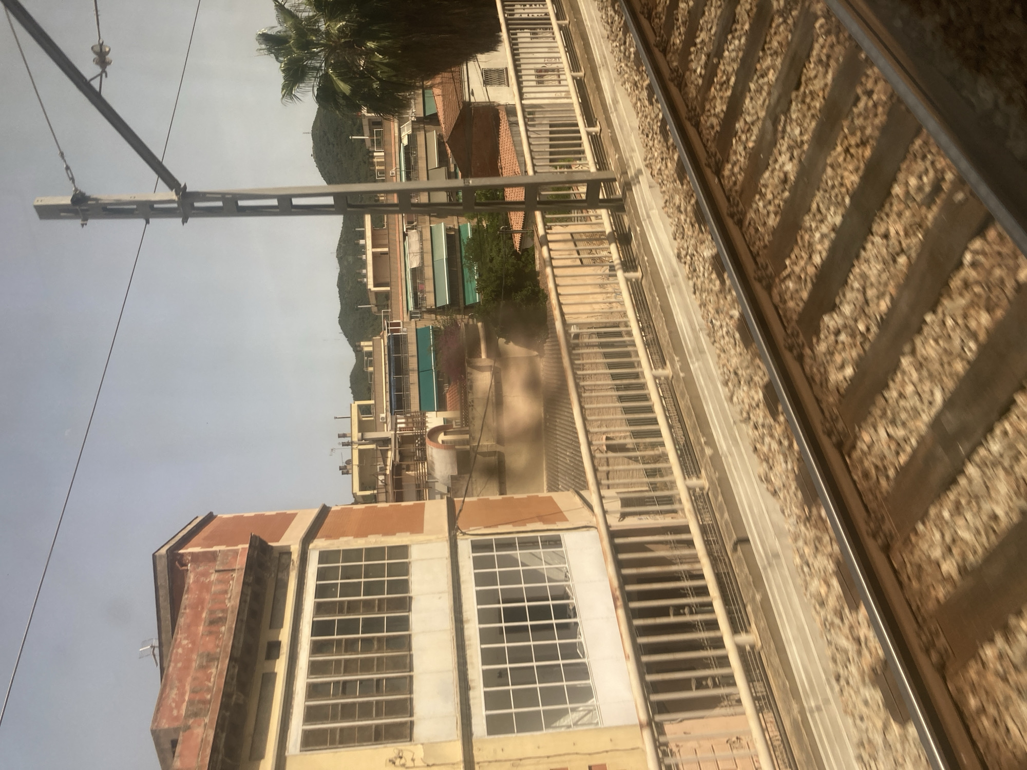 The image captures a view from a moving train in Montcada i Reixac, Spain. The foreground shows train tracks with ballast and wooden sleepers. In the midground, a bridge railing separates the tracks from a built-up area. To the left, a building with a yellow lower facade and an orange upper facade features large windows with white frames. Behind this, several other buildings are visible, some with balconies and awnings. A tall, grey utility pole with overhead wires stands prominently to the right of the bridge. Further in the background, green hills and a palm tree are discernible against a hazy sky.