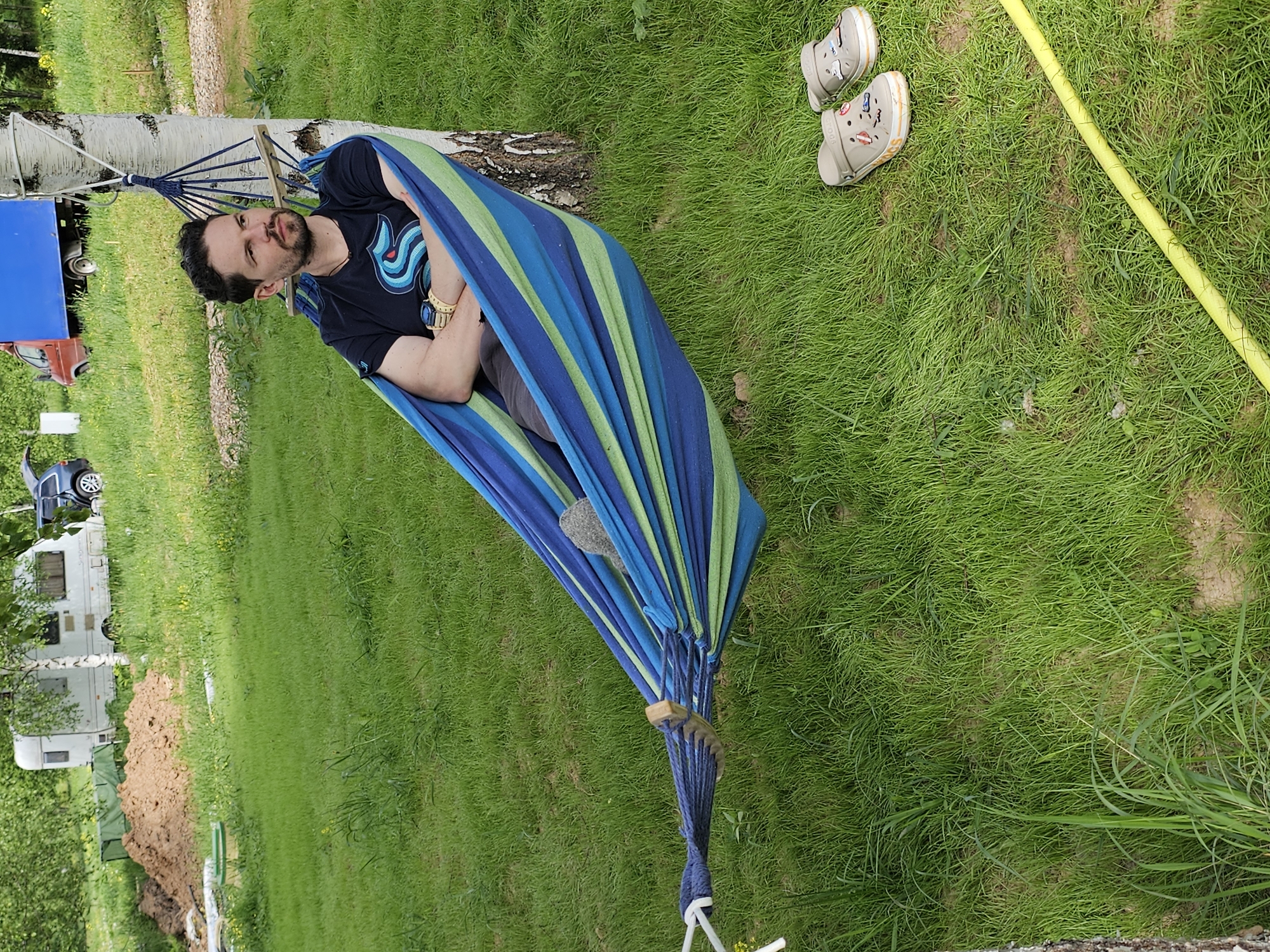 A man is relaxing in a blue and green striped hammock suspended between two trees in a grassy area, likely in Мышкино, Russia. He is wearing a black T-shirt with a distinctive design, grey shorts, and a gold watch. His footwear, a pair of beige sandals adorned with small embellishments, are placed on the grass beside the hammock. A yellow hose lies near the sandals. In the background, there is a trailer, a blue car, and a partially visible red car. A pile of freshly dug earth and construction debris are visible near the trailer. The scene suggests a casual, outdoor leisure setting.