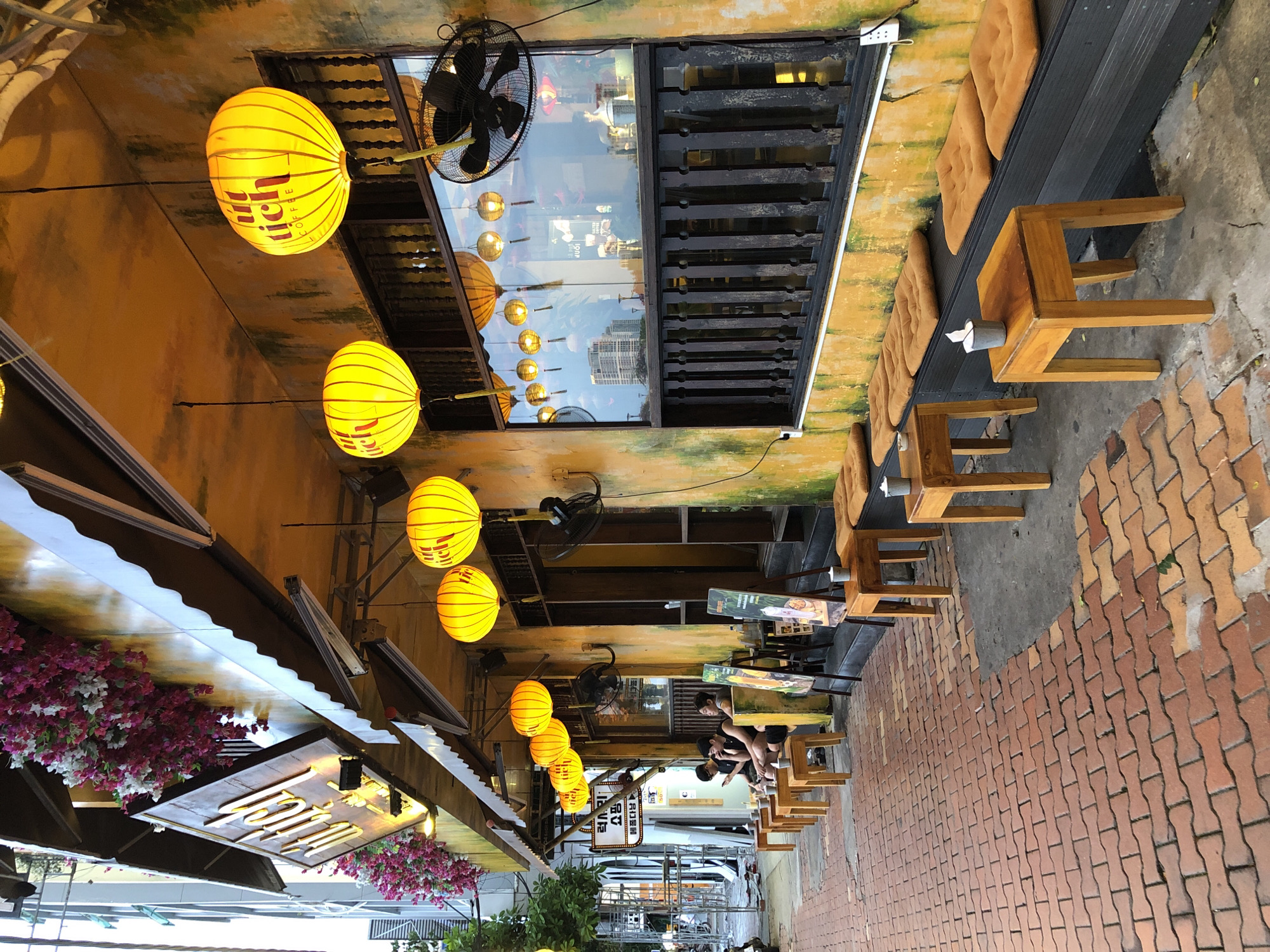 The image depicts an outdoor seating area of a cafe named "UT TIC COFFEE" located in the city of Đà Nẵng, Vietnam. The facade of the cafe is painted a vibrant yellow with some green moss-like staining, creating a weathered appearance. A white awning extends over the seating area, and it is decorated with cascading pink and white flowers. Several round, yellow paper lanterns with the cafe's logo are suspended from the awning and under the roofline.
The seating area consists of low wooden tables and benches with orange cushions. A brick-paved walkway leads up to the seating area. In the background, through a large window, the interior of the cafe is visible, with reflections showing buildings and the sky. A black industrial-style fan is mounted on the wall near the window.
A sign with Vietnamese text is visible on the left side of the cafe, and several patrons are seated at the tables, seemingly engaged in conversation or looking at their phones. The overall atmosphere suggests a casual and inviting cafe environment in a bustling city.