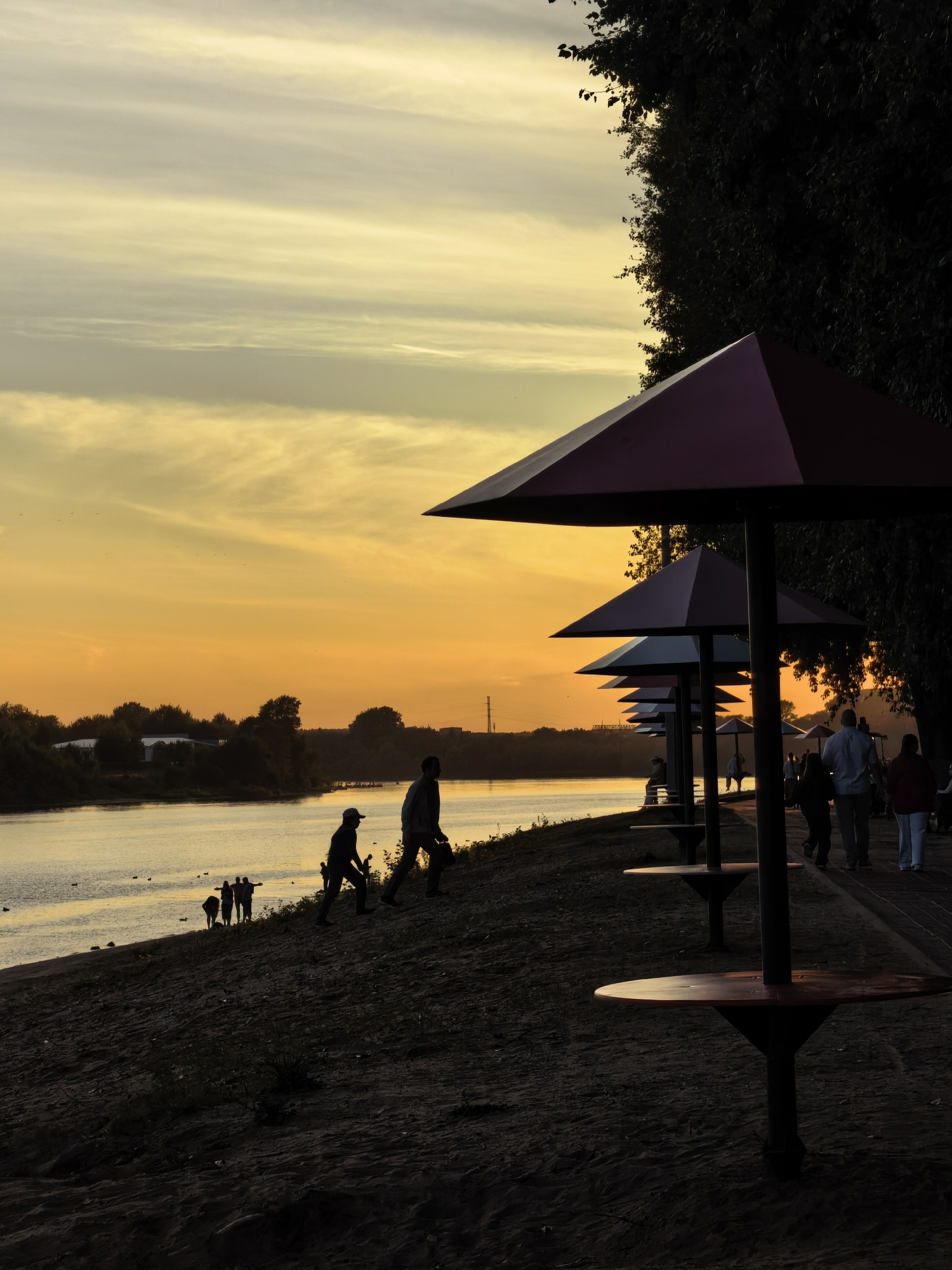 The image depicts a sunset scene along the bank of the Volga River in Tver, Russia. The sky is a gradient of yellow and orange, with wispy clouds. In the foreground, a sandy embankment leads to the river. A row of covered picnic tables, silhouetted against the sky, are positioned on a paved walkway parallel to the river. Several people are walking along the walkway, their figures dark silhouettes. On the embankment nearer to the river, a group of individuals, also silhouetted, are visible. The river itself is calm, reflecting the colors of the sky, and a few ducks are swimming on its surface. The overall atmosphere is peaceful and serene, characteristic of an evening by the water.