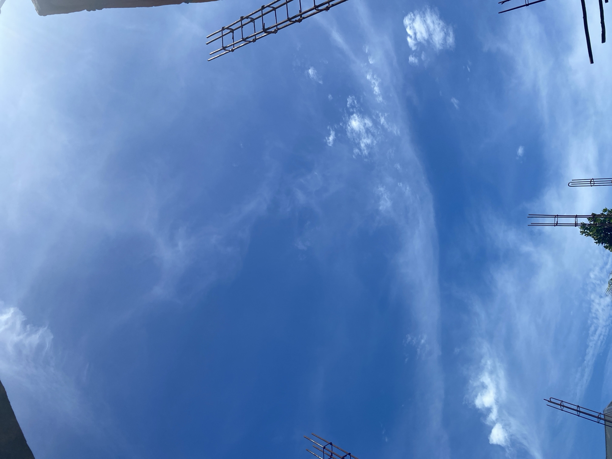 The image is a low-angle shot looking up at a bright blue sky with scattered white clouds. In the foreground, several rebar cages, common in concrete construction, are visible protruding upwards. These structures suggest an ongoing construction site. The lighting indicates it is daytime, likely sunny with some haze from the thin clouds. The top right of the image shows a dark, rectangular object, possibly a building or scaffolding, partially obscuring the sky. The lower part of the frame reveals more rebar structures and some greenery, possibly trees. The scene is consistent with a construction site in a tropical climate, such as Banda Aceh, Indonesia, given the clear, bright sky and the presence of construction materials. There are no people visible in the frame.