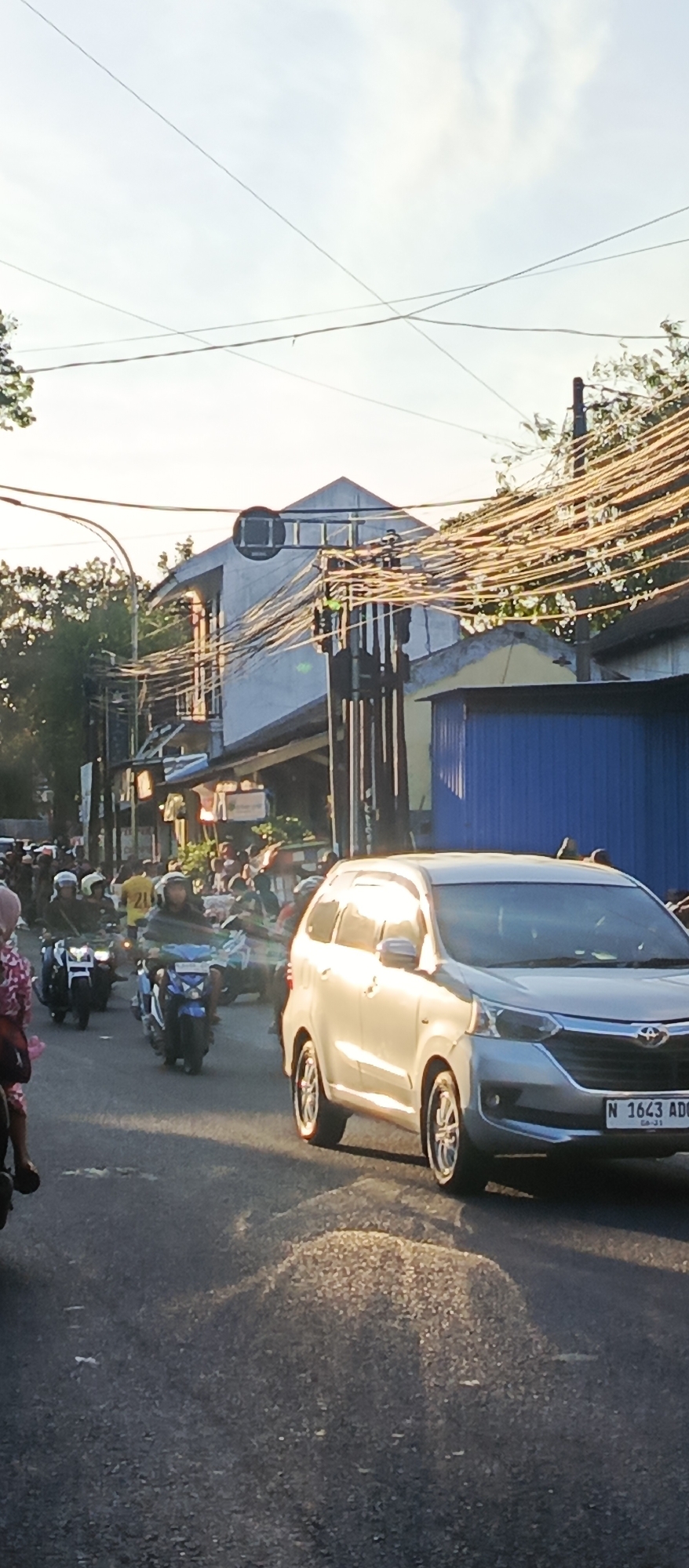 The image captures a street scene in Malang, Indonesia, under what appears to be late afternoon sunlight. A silver car, with license plate N 1643 ADC, is prominently featured in the foreground, traveling on an asphalt road. The car's reflection is visible on the road surface, creating a warm, dappled effect due to the low angle of the sun. In the background, a busy street unfolds with numerous motorcycles and their riders. The motorcycles are of various colors, including blue and white. Some individuals are wearing helmets, and one person on the left side of the frame is wearing a pink and white patterned outfit. The street is lined with buildings, some of which appear to be shops or residences with awnings and signs. A significant feature of the urban environment is the dense network of electrical wires and poles, crisscrossing overhead and creating a somewhat cluttered visual impression. Trees with lush green foliage are visible behind the buildings, suggesting a vegetated area within the city. The overall atmosphere is one of a bustling urban environment, indicative of daily life and traffic in a Southeast Asian city.