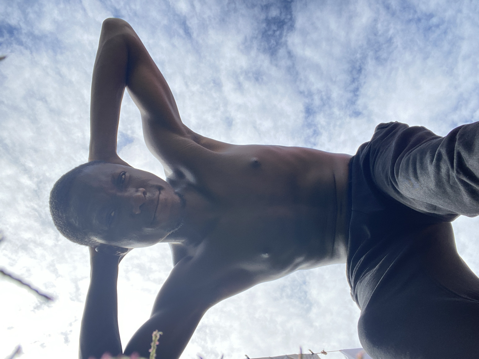 The image is a low-angle, outdoor shot of a shirtless Black man wearing dark pants. He is looking up towards the sky, with one arm raised and bent behind his head, and the other arm bent and resting on his head. The background is dominated by a cloudy sky, suggesting it is daytime. The man's expression appears relaxed or contemplative. The location is identified as Bukuru, Nigeria. There is no visible text in the image.