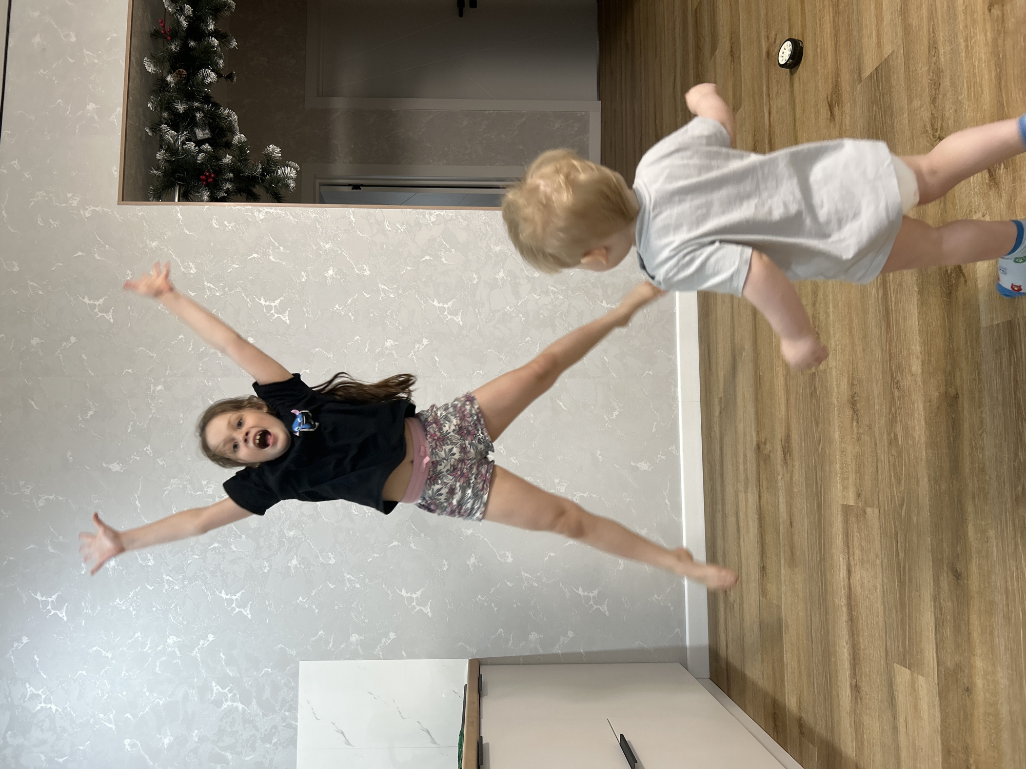 In an indoor environment, a girl in a black shirt and floral shorts is airborne with arms outstretched and legs spread. A toddler in a light-colored shirt is being held aloft by the girl. A Christmas tree is visible in the background, and a clock is affixed to the wall. The location is possibly in Chaikovsky, Russia.
