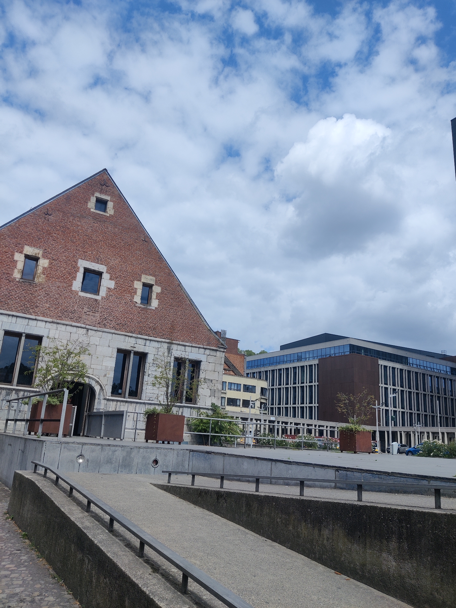 This image depicts the exterior of the Grand Curtius Museum in Liège, Belgium. The museum complex features a combination of historical and modern architecture. In the foreground, a concrete ramp with metal railings leads towards the entrance of the historical building. This older section of the museum is characterized by its red brick facade, stone detailing around the windows, and a steep gabled roof with small, square windows. Several potted plants are placed along the upper level of this historical section.
To the right of the historical building, a modern, multi-story building with a glass facade and dark vertical elements stands prominently. This contemporary structure houses offices or other facilities and contrasts sharply with the older architecture. The background reveals a blue sky with scattered white clouds, suggesting fair weather. The scene appears to be a public space, possibly a plaza or courtyard, situated within an urban environment. There are no people visible in the image.