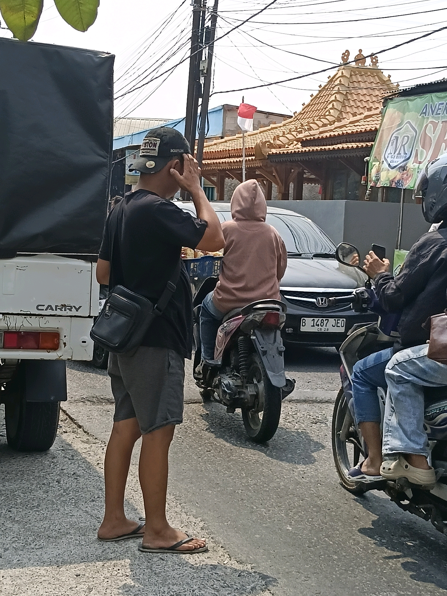 The image depicts a street scene in Belimbing, Indonesia, with a focus on pedestrians and vehicles at what appears to be an intersection. In the foreground, a man wearing a black t-shirt, grey shorts, and flip-flops stands with his hand to his ear, looking to his right. He carries a black crossbody bag. To his left, a portion of a white truck with the word "CARRY" visible on its side is present.
Behind the man, a person wearing a light brown hooded sweatshirt is seated on a motorcycle. Another motorcycle is to the right of this one, with a rider wearing a black jacket and jeans, holding a smartphone. Both motorcycles are stopped, suggesting traffic or a pause in movement.
In the background, a building with a distinctive tiled, ornate roof is visible, along with a red and white flag on a pole. A banner with green and white text, including "ANER" and "AR GRIBRIO," is also present. Overhead, a complex network of electrical wires is strung across poles, typical of urban environments. The ground is paved with asphalt and appears somewhat weathered. The overall atmosphere suggests a typical day on a busy Indonesian street.
