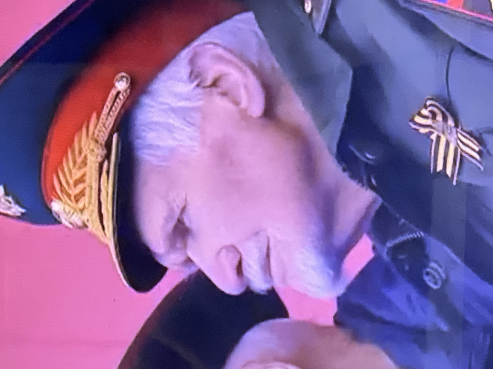 A close-up shows a person in a military uniform and hat. The uniform is dark green with a gold and black ribbon pinned to the lapel. The hat is dark with a red band and gold trim. The person has white hair and their face is obscured by the angle of the shot. The background is a solid pink color. The location is potentially Chaikovsky, Russia.