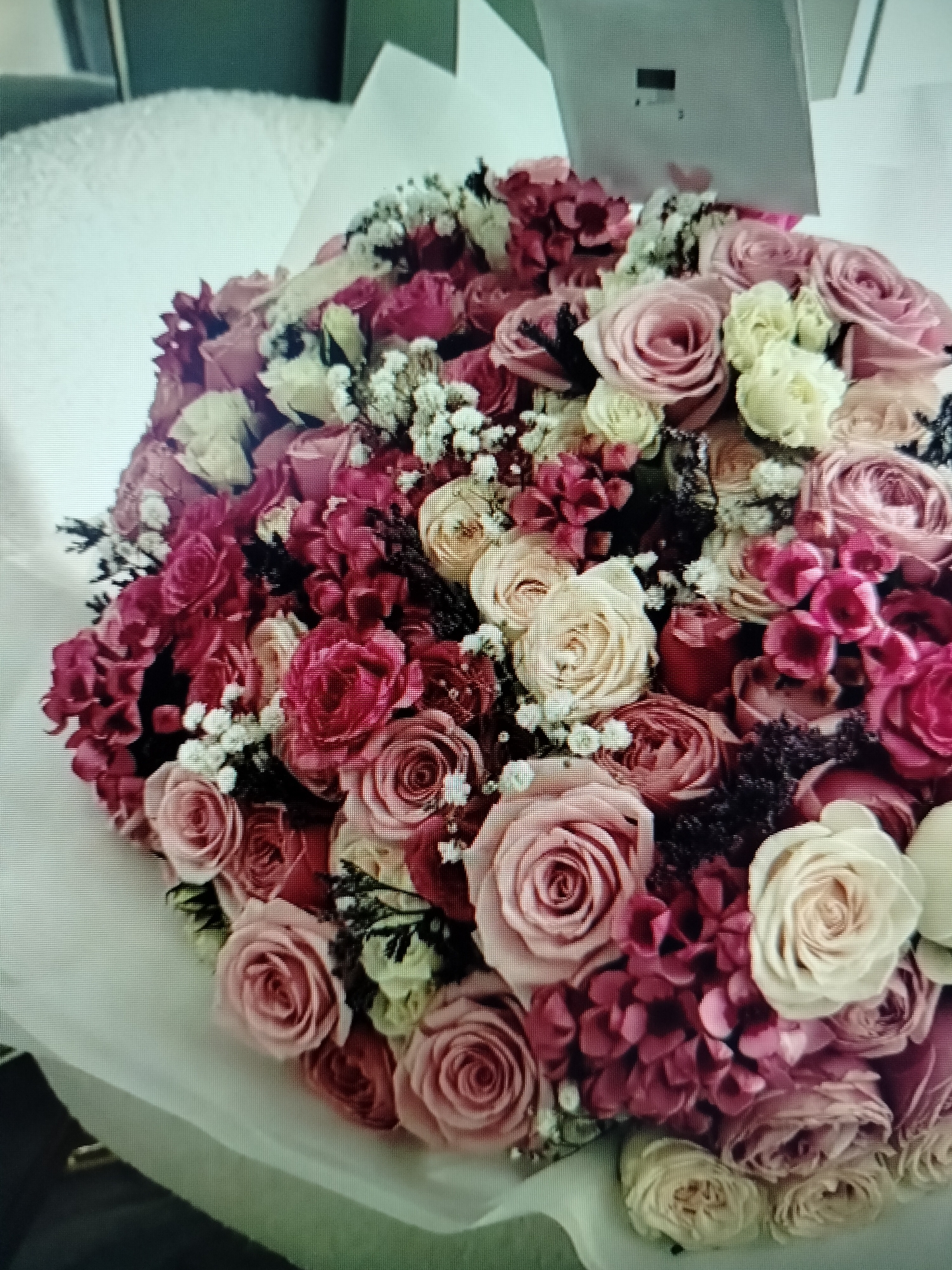 A close-up, top-down shot shows a large, round bouquet of flowers. The bouquet is arranged in a dense cluster and is partially wrapped in white paper. The flowers appear to be primarily roses and hydrangeas in various shades of pink, red, and cream, with some small white filler flowers interspersed throughout. The background is blurry and appears to be a light-colored, textured surface, possibly a tablecloth or sofa. There are no people, actions, or interactions visible in the image, and no elements suggest a specific location in Yashkino, Russia.