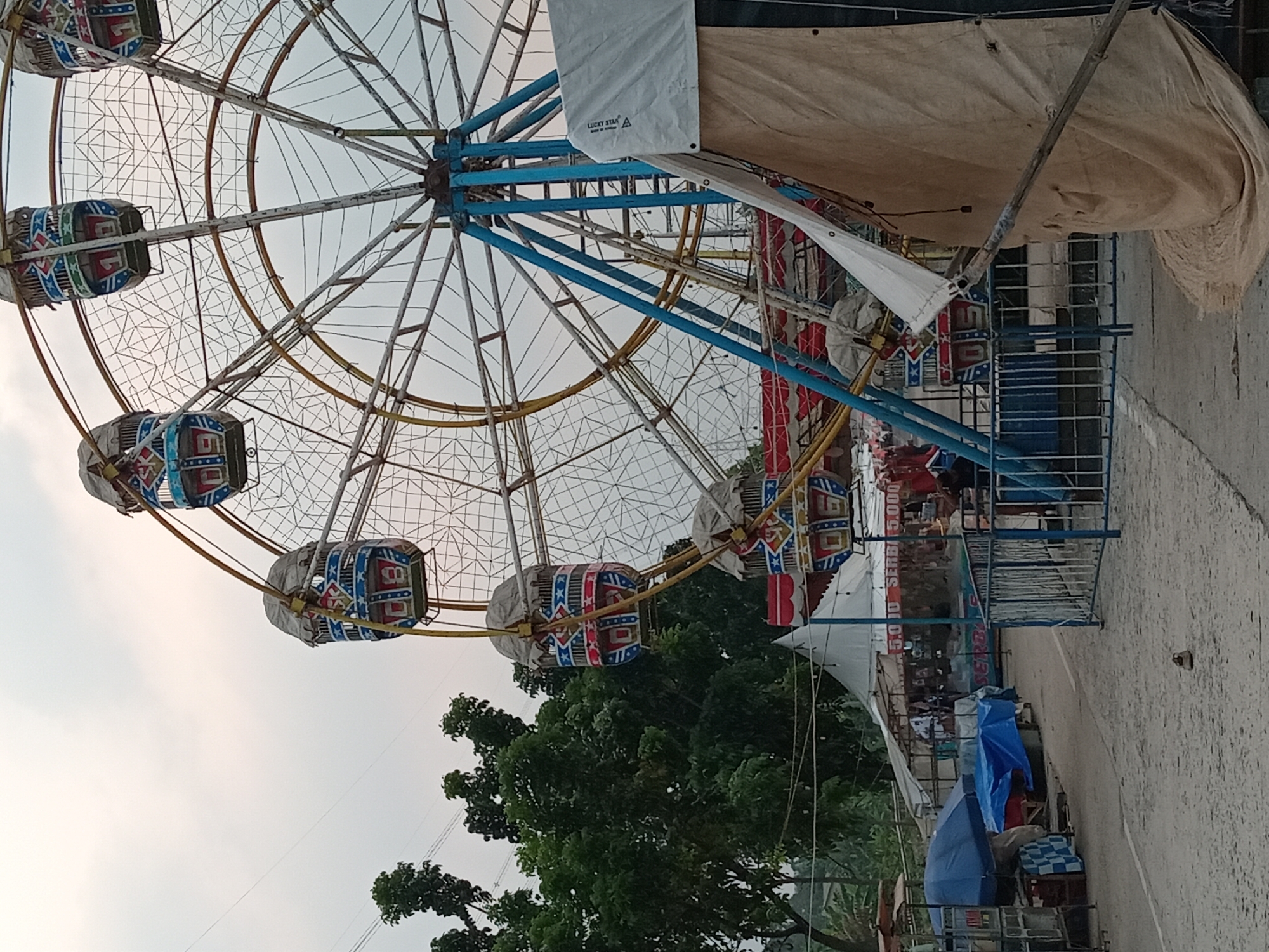 The image captures a Ferris wheel at a fairground in Depok, Indonesia. The Ferris wheel, with its multi-colored cabins, dominates the upper half of the frame, while the lower portion shows a path lined with tents and stalls. Several cabins on the Ferris wheel are partially covered with fabric. A blue metal fence separates the path from the base of the Ferris wheel. Tents, some white and some beige, are set up behind the fence, with visible signs, one of which reads "5.000 SERBU 5.000". Trees and foliage are present in the background, suggesting an outdoor setting. The ground is paved with a rough surface. A blue umbrella is visible near the stalls. A lone sandal rests on the path. The sky is overcast.