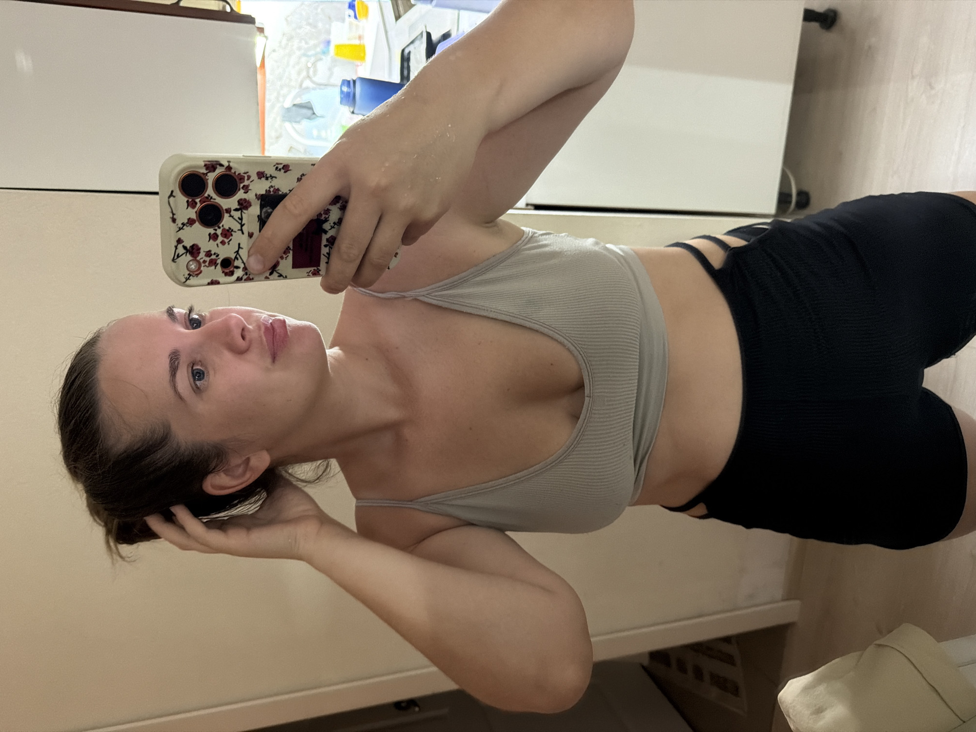A young woman with brown hair pulled back in a messy bun and blue eyes takes a selfie in a mirror. She is wearing a light grey ribbed tank top and black ribbed shorts with cutouts on the sides. Her left hand is touching her hair, and her right hand holds a phone with a floral case. She appears to be in a bathroom or kitchen, with white cabinets and a tiled floor visible in the background. There are also some blue and yellow containers on a shelf. The image is well-lit and sharp, with the woman in focus.
