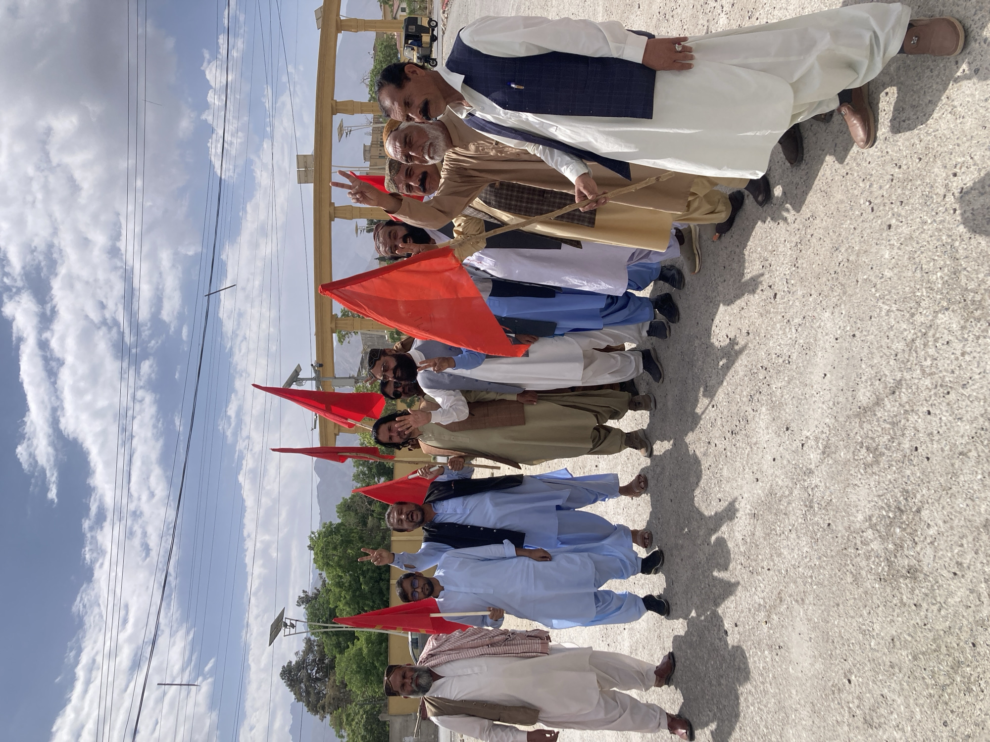 A group of approximately ten to twelve men is captured in an outdoor setting, arranged in a diagonal line from the foreground to the background. Most are looking towards the camera, smiling, and several are making peace signs with their fingers, suggesting a positive and possibly celebratory or peaceful atmosphere.
The men are dressed in traditional South Asian attire, primarily white or light-colored shalwar kameez. Some wear dark or light-colored waistcoats over their kameez, while others have shawls or turbans. Several individuals in the mid-ground and foreground are holding plain red flags on wooden poles, indicating a unified group, possibly for a procession, rally, or gathering.
The scene appears to be a street or open area with a rough, light-colored ground surface. Overhead, numerous electrical wires crisscross against a bright sky with scattered white clouds. In the background, green trees and light-colored buildings with prominent columns are visible, along with poles equipped with solar panels. Distant mountains frame the horizon. The strong sunlight casts distinct shadows of the men on the ground, indicating it is daytime, likely midday or early afternoon. This scene, set in Quetta City Tehsil, Pakistan, portrays a community event or demonstration. No discernible text is visible on the flags or other objects.