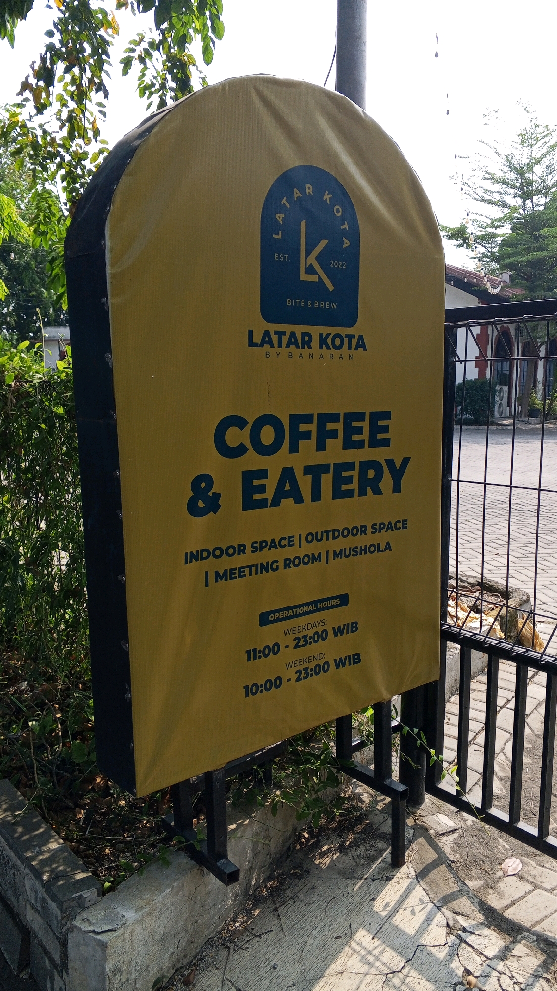 A yellow banner sign with dark blue text is displayed on a black metal stand. The sign is located outdoors, with greenery and trees in the background. The sign has a logo at the top which reads "LATAR KOTA EST. 2022 BITE & BREW". Below the logo, it says "LATAR KOTA BY BANARAN COFFEE & EATERY". Further down, it lists features: "INDOOR SPACE | OUTDOOR SPACE | MEETING ROOM | MUSHOLLA". Finally, it states "OPERATIONAL HOURS WEEKDAYS: 11:00 - 23:00 WIB WEEKEND: 10:00 - 23:00 WIB". The sign is positioned in front of a black metal fence and a paved area. The context of Semarang, Indonesia is inferred from the provided text on the sign.