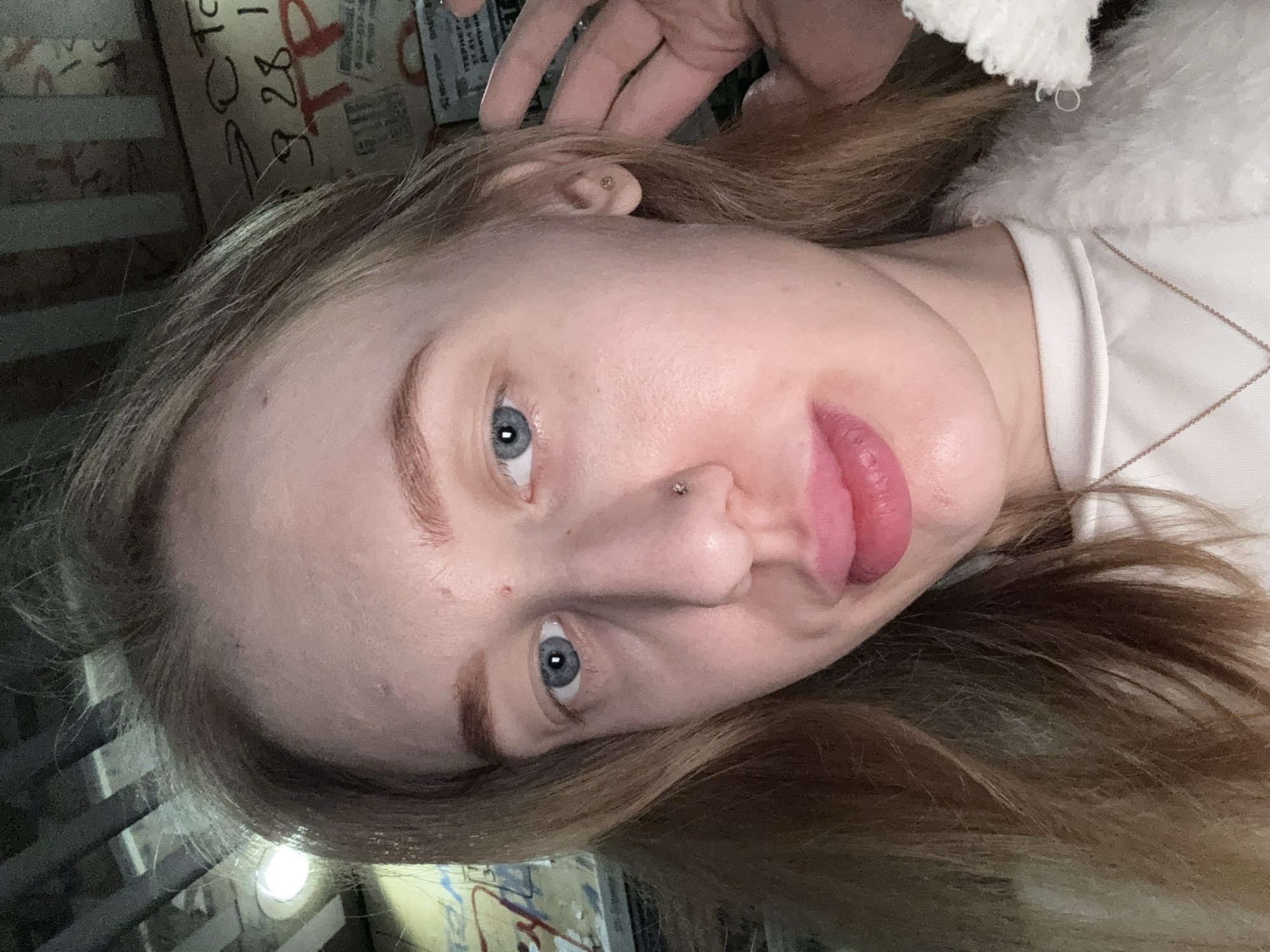 A close-up, angled selfie depicts a fair-skinned female with light brown hair and blue eyes. She has a nose piercing and an ear piercing. She is wearing a white shirt, partially obscured by a white fuzzy garment. The background appears to be a blurred interior space with indistinct objects and text visible. It is not possible to confirm the location as Koysugskiy, Russia based on visual information in the image.