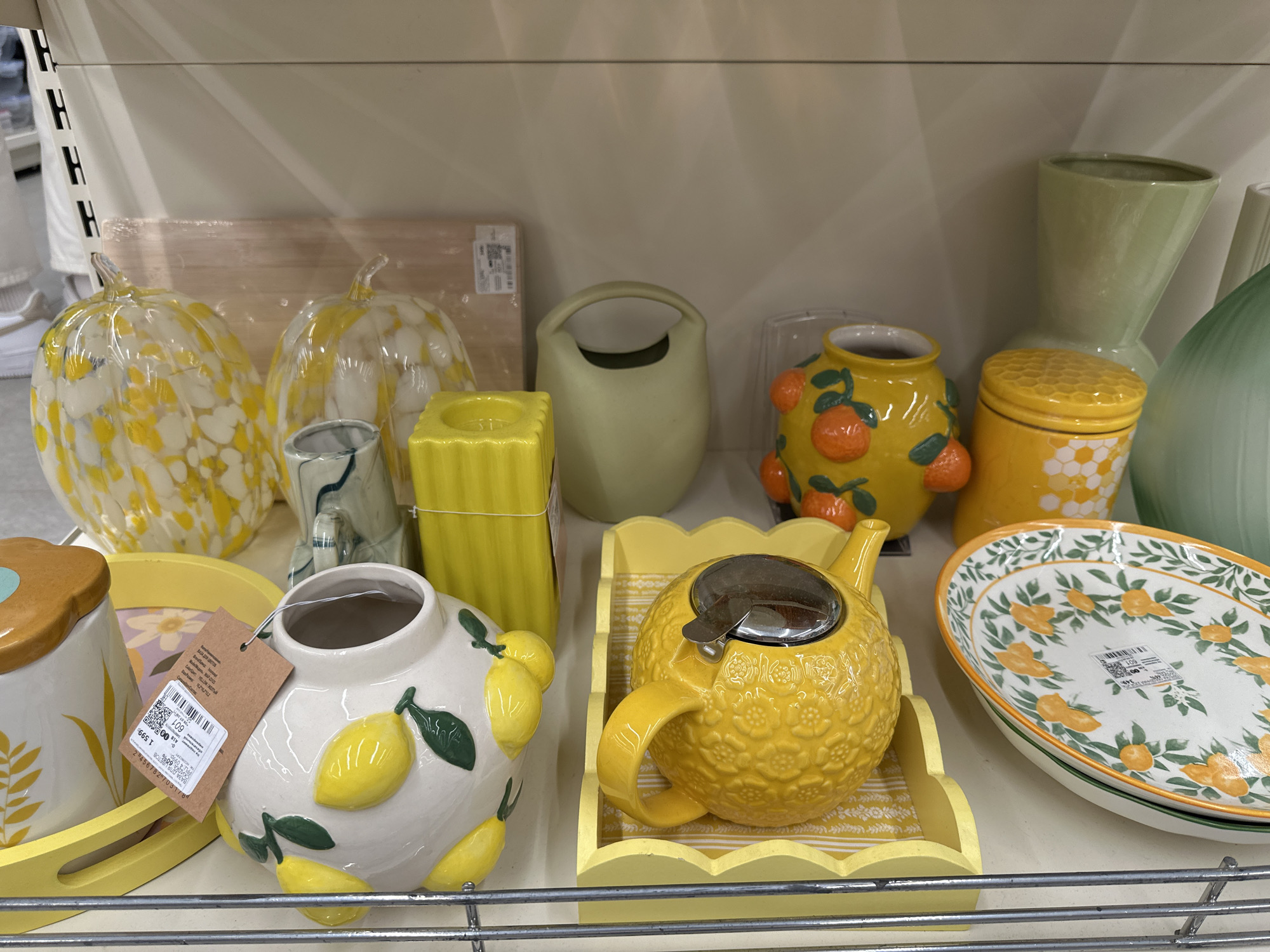 On a shelf in a retail store, likely in Samara, Russia, several decorative ceramic and glass items are displayed. The items are arranged in rows and include two speckled glass pumpkins in yellow and white, a green ceramic jug with a handle, a yellow ribbed candle holder, a white ceramic vase decorated with lemons and leaves, and a yellow teapot with a textured pattern, sitting on a yellow scalloped tray. To the right, a green vase and a large oval plate with an orange fruit and green leaf pattern are visible. The items are placed on a light-colored shelf against a plain background. There are no people in the image.