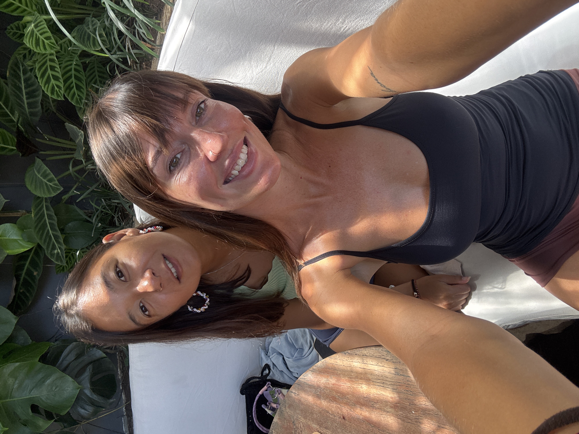 Two women are in a selfie taken outdoors. The woman in the foreground wears a black tank top. The woman in the background is wearing flower-like earrings. The background includes various plants. The image was taken in Kuta Selatan, Indonesia, as indicated by the location data. A black purse and light blue denim jacket are near the woman in the background. They are both laying on a white mattress.