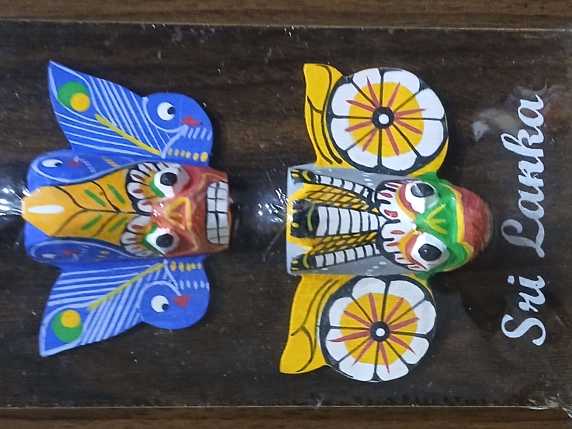The image displays a pair of painted wooden masks, mounted on a dark wood backing labeled "Sri Lanka" in white script. The masks are brightly colored and stylized. The mask on the left features blue "wings" decorated with stylized peacock feather patterns, a yellow and red face, and white teeth. The mask on the right has yellow "wings" resembling flower petals, a gray and white body, and a green and red face. The masks are connected by clear plastic wrap and placed in the center of the wood. The setting or location cannot be determined from the provided image.