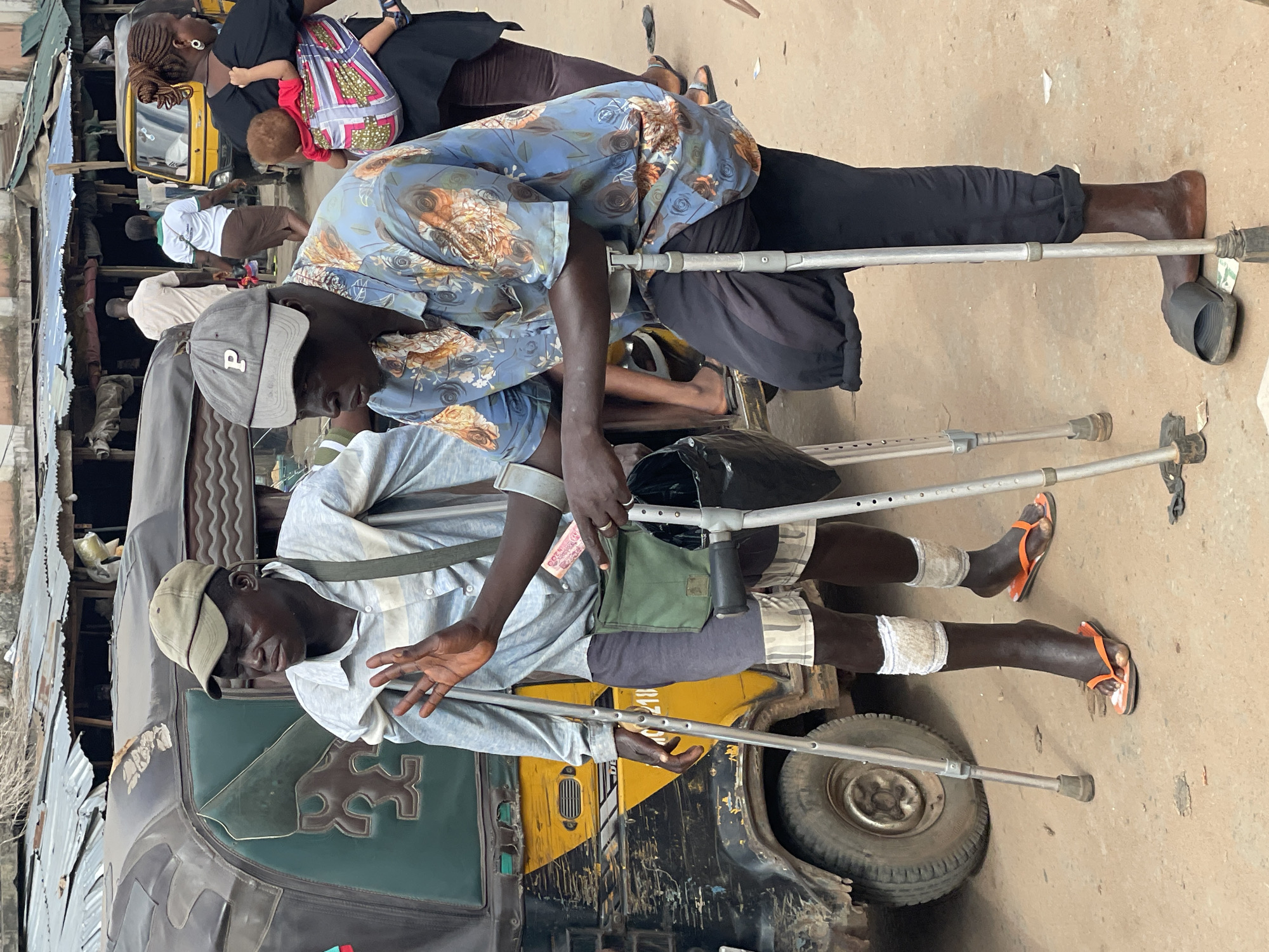 The image depicts two men, both using crutches, sitting on what appears to be a vehicle, possibly a tricycle taxi, in Nkpor, Nigeria. The man in the foreground is wearing a light-colored shirt, camouflage shorts, and a cap, with bandages on his legs and orange flip-flops. He is holding crutches. The man behind him is wearing a floral-patterned shirt, dark pants, and a gray cap, also holding crutches. A black bag is near them. Other people are visible in the background, including a woman carrying a child. The vehicle they are sitting on is partially painted green and yellow and shows signs of wear. The surrounding environment suggests an urban setting with buildings and other vehicles.