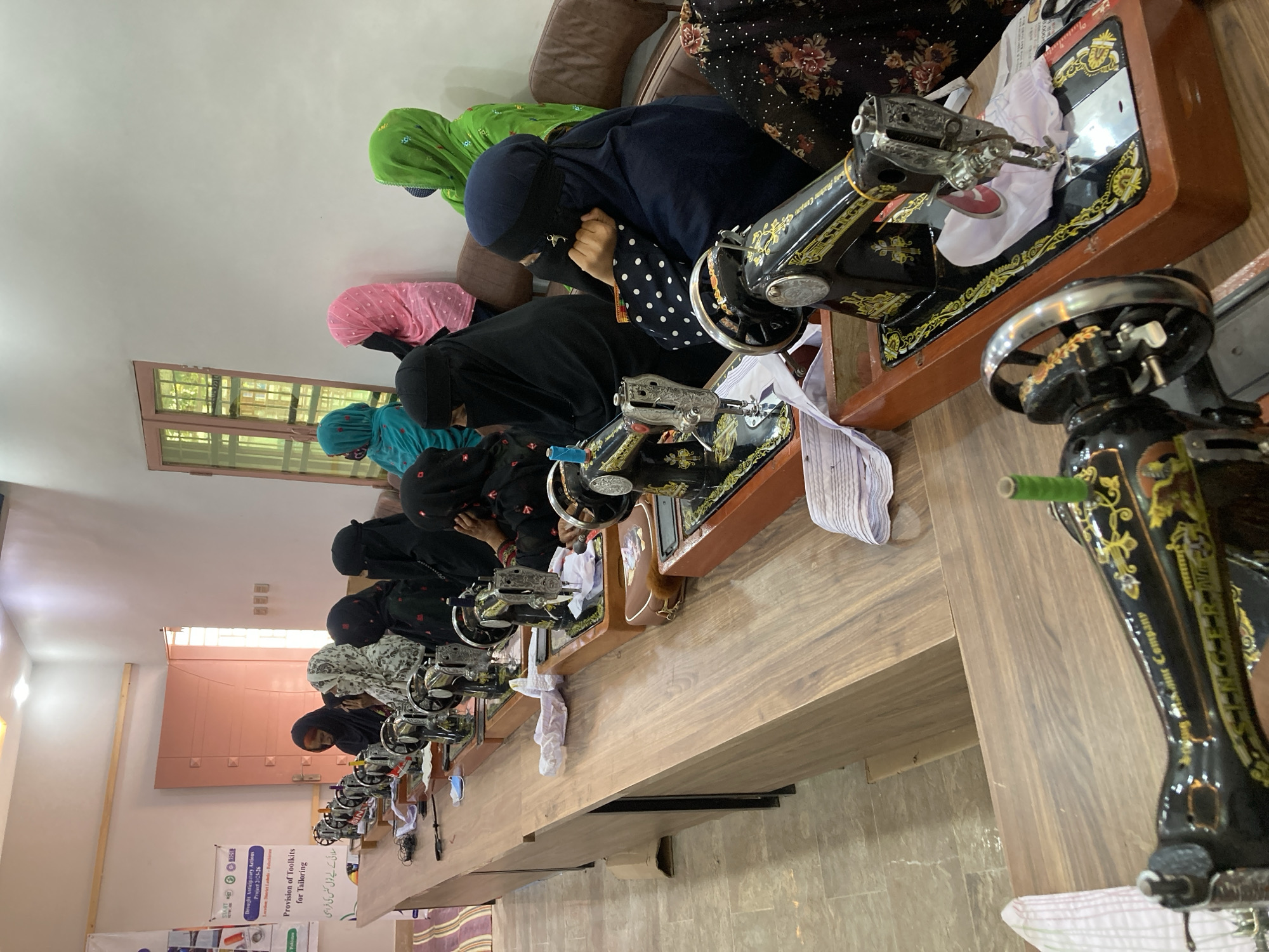 A group of women, all wearing hijabs and some in niqabs, sit side-by-side at a long wooden table. Each woman operates a vintage, black Singer sewing machine. White fabric is placed on the sewing machine tables in front of each woman. The women are likely taking a tailoring class. A sign in Urdu and English on the far left reads "Provision of Toolkits for Tailoring" and mentions "Drought Anticipatory Actions, Project 2-25-26" suggesting this is part of an aid project. The location is Rāwāni, Pakistan. A window is visible in the background. A brown couch is placed behind the women.