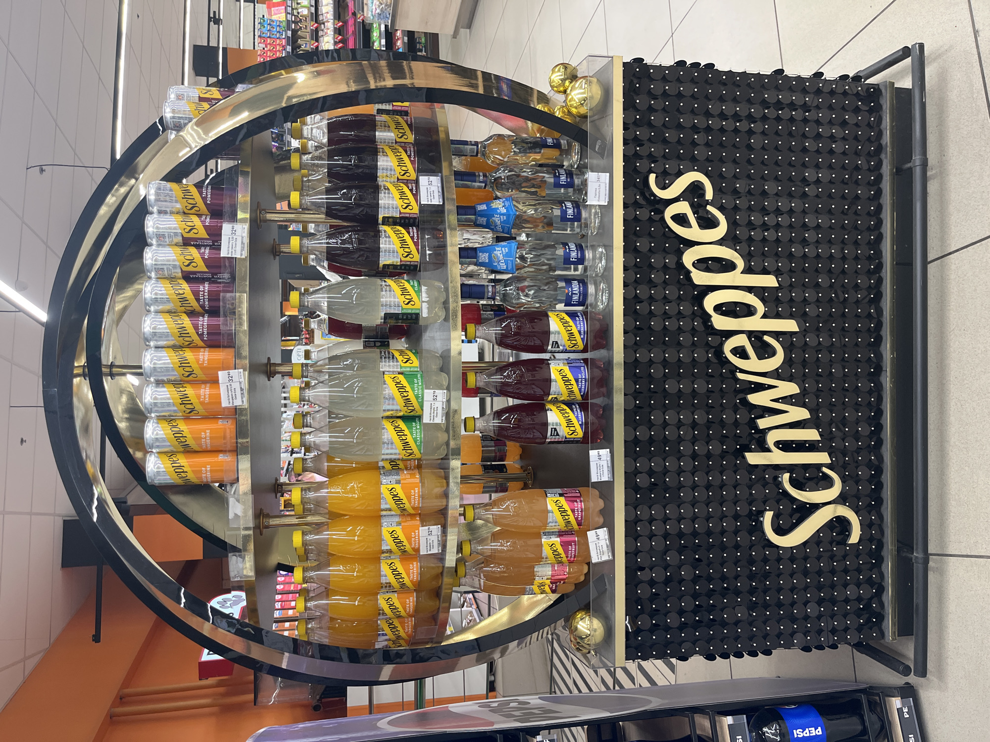 A multi-tiered display rack featuring Schweppes beverages is centrally located in a brightly lit retail environment, possibly a supermarket or convenience store. The rack has a distinctive circular, gold-colored frame. On the top shelf, cans of Schweppes are arranged, showcasing flavors such as Tangerine and Pomegranate. The middle shelf displays bottles of Schweppes drinks, including Classic Mojito, and on the bottom shelf, there are bottles of Schweppes Blueberry Mojito and Finlandia Vodka. The base of the display is adorned with black sequins and prominently displays the Schweppes logo in gold lettering. Golden decorative spheres are placed on the bottom shelf. To the left of the display, a part of a Pepsi advertisement is visible. The flooring appears to be tiled. The overall scene suggests a product promotional setup. The location is in Kharkiv, Ukraine, as indicated by the presence of "Finlandia" which is a product commonly sold there.