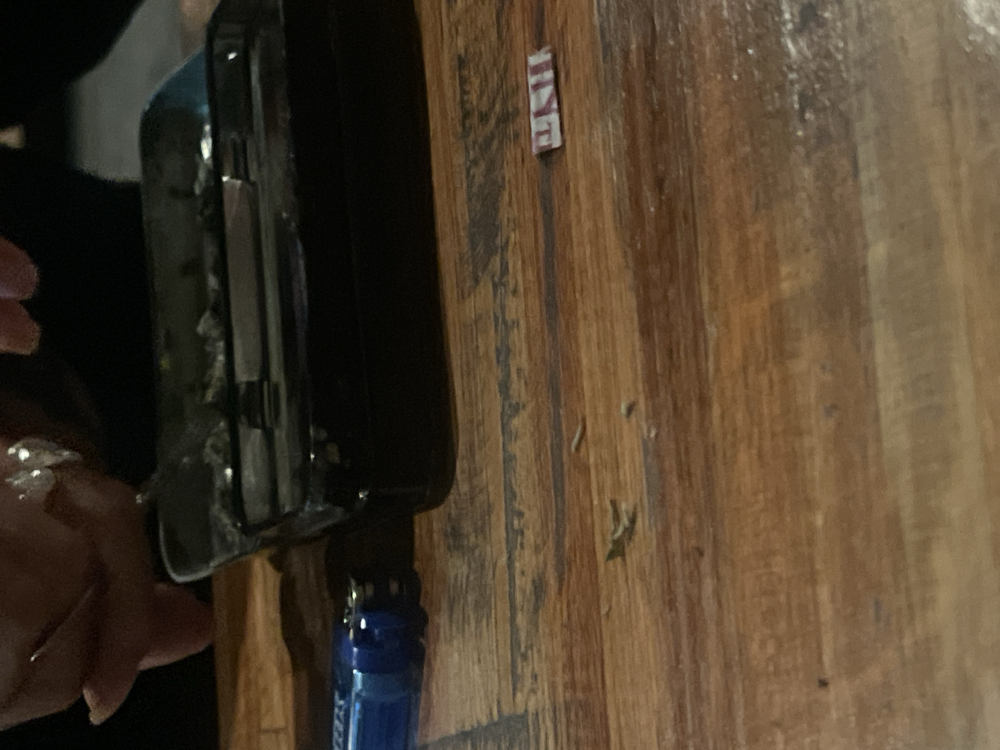 A black smartphone is partially held by a hand with visible rings on a wooden surface. An item with "MINE" printed on it is on the surface. A blue bottle-like object is partially visible attached to the smartphone. The location and any interactions cannot be determined from the image.