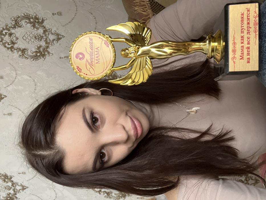 A young adult female is depicted indoors in Chaikovsky, Russia, holding a golden trophy. The woman, with long dark brown hair parted down the middle, brown eyes, a small nose piercing, and light makeup, looks directly at the camera with a subtle smile. She wears a light beige off-shoulder top and a silver-colored necklace with a small winged pendant. A gold hoop earring is visible in her right ear. The trophy features a golden winged female figure holding a circular emblem. The emblem has a reddish-pink background with a gold border and displays a mother and child silhouette icon, along with the Russian text "Любимая МАМА" (Beloved MOM). The trophy rests on a dark rectangular base, which has a light-colored plaque bearing the Russian inscription "Мама как пуговка: на ней все держится!" (Mom is like a button: everything holds on her!) in red lettering. The background consists of light-colored wallpaper with an ornate gold and beige damask pattern and a section of a patterned fabric (possibly a blanket or sofa throw) to the right.