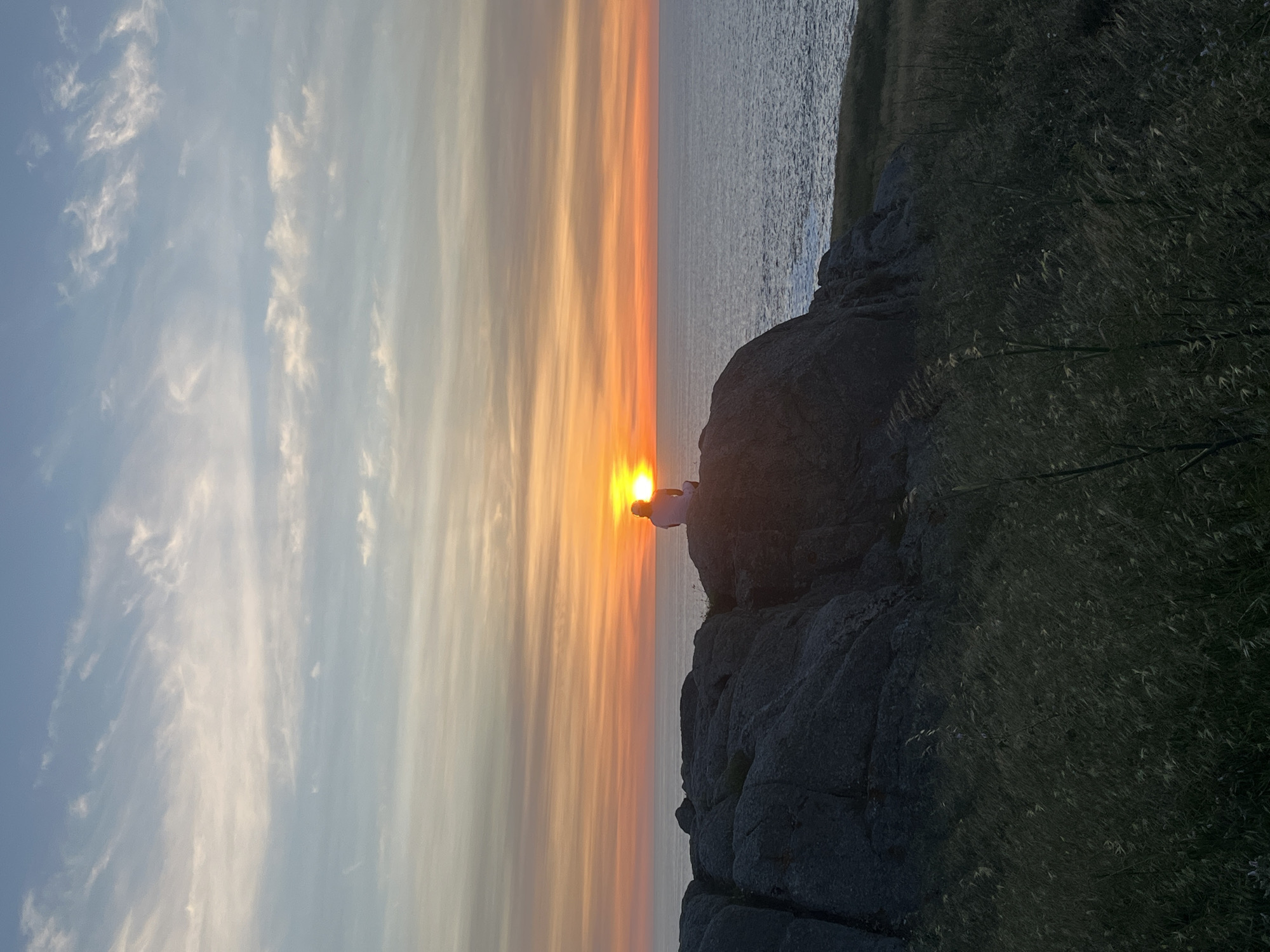 The image shows a sunset view, with the sun positioned near the horizon line over water. The sky displays a gradient of colors, from light blue with wispy clouds at the top to orange and yellow near the horizon. A rocky outcrop with vegetation is in the foreground. A figure is visible on top of the rocks near the sun, silhouetted against the bright light. The location is near the ocean, but the specific city in Spain is unknown.
