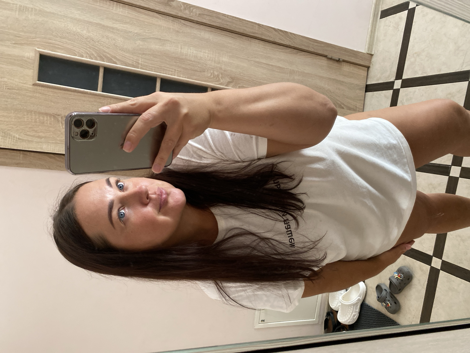 A woman with long, dark brown hair and blue eyes is taking a mirror selfie. She is wearing a white t-shirt. In the reflection, a wooden door is visible in the background, as is a checkered floor with white and dark grey tiles. To the left, a shelf with various shoes is visible, including white platform slides and grey toddler-sized Crocs. The scene appears to be an indoor setting, possibly a home or hotel room, and there is no indication of it being in Sochi, Russia.