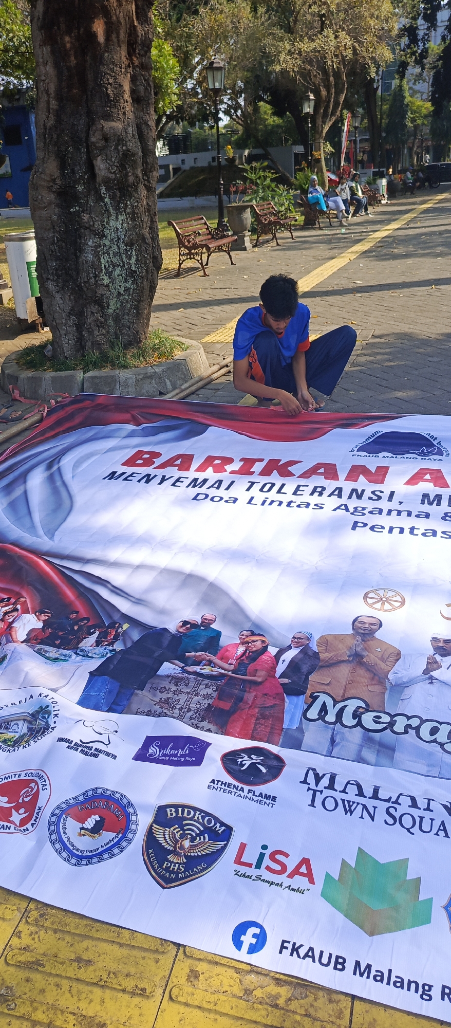 The image depicts a young male, seemingly in his late teens or early twenties, kneeling on the ground and working on a large banner laid out on a public sidewalk. The banner features bold red and white lettering with Indonesian text, including "BARIKAN A," "MENYEMAI TOLERANSI, ME," and "Doa Lintas Agama & Pentas." The banner is also adorned with various logos and a photographic collage of people from different religious backgrounds interacting, suggesting an event promoting religious tolerance. In the background, park benches, trees, and street lamps are visible, indicating an outdoor public space, likely a park or pedestrian area in the city of Malang, Indonesia. The overall scene suggests preparation for a community event.